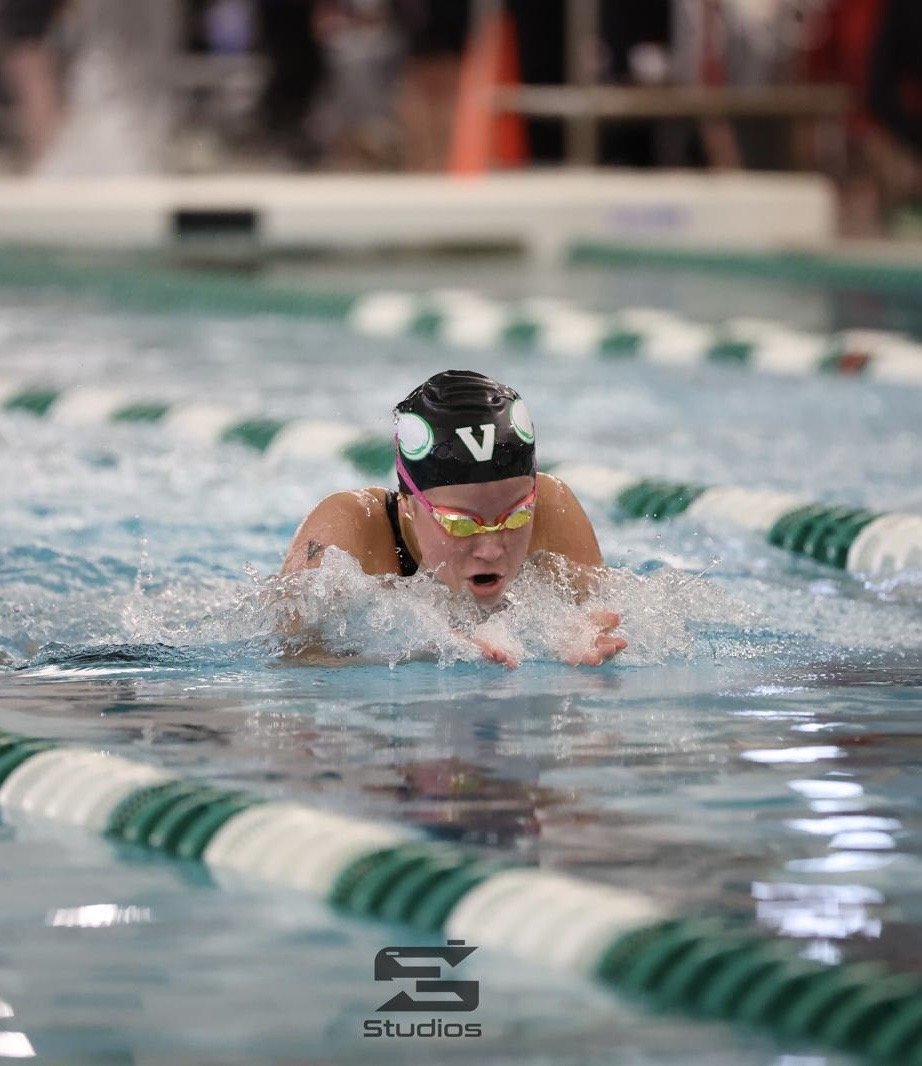 A female swimmer wearing a black swim cap with a white letter V, goggles, and a black swimsuit is competing in a swimming race in an indoor pool. She is performing the breaststroke, with her face submerged and arms moving forward. The pool has green and white lane dividers, and the background shows some blurred equipment and racks.