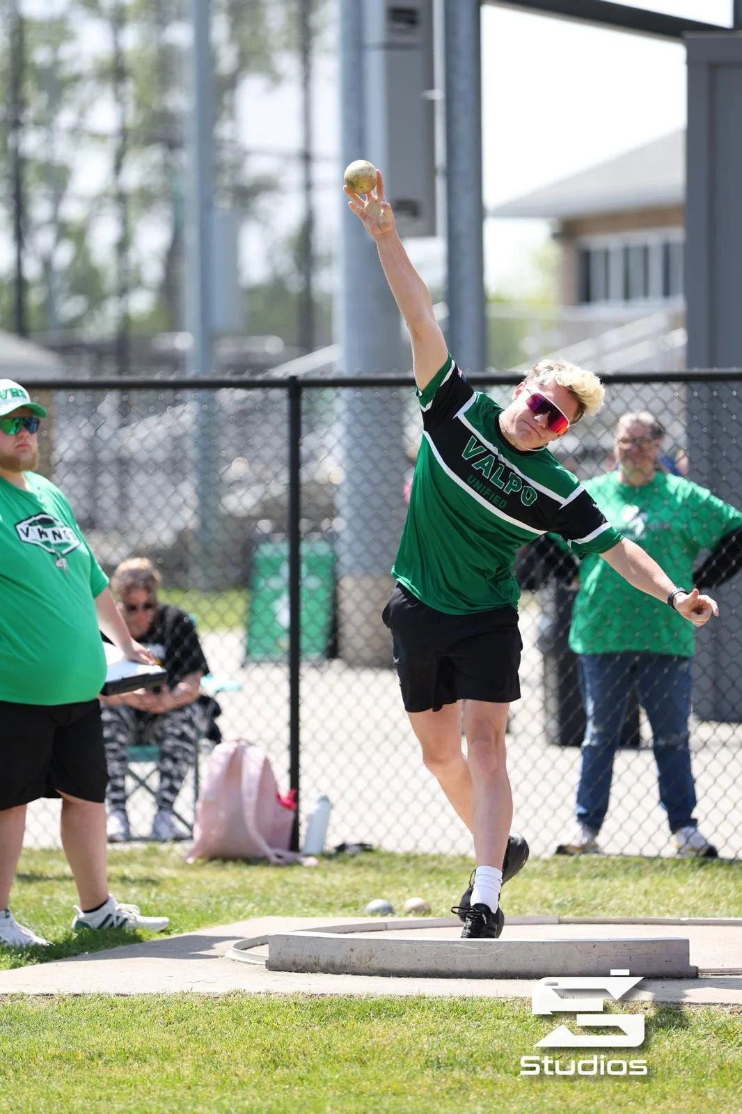 A young man in green sports attire and sunglasses throws a shot put with other spectators watching behind a fence during a sunny outdoor event.