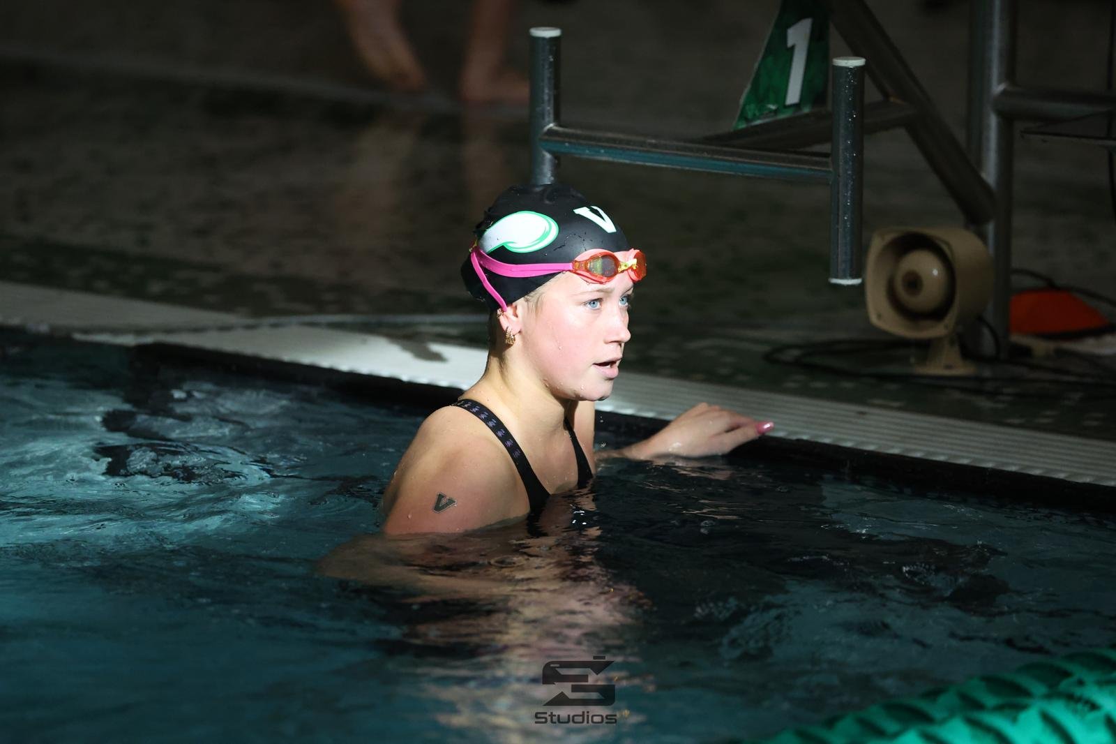 Female swimmer wearing a black swim cap with a white and green design, pink goggles on her forehead, and a black swimsuit, standing in a swimming pool holding on to the pool edge.