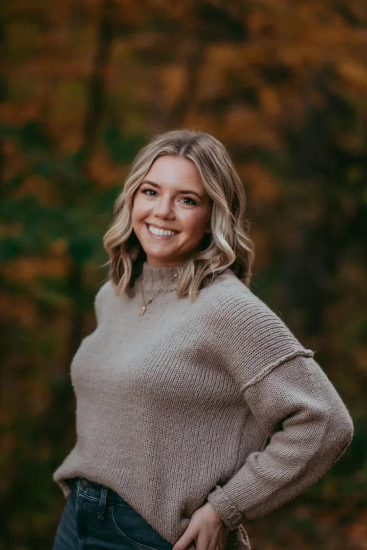 A young woman with blonde, wavy hair smiling outdoors during autumn, wearing a beige sweater and blue jeans.