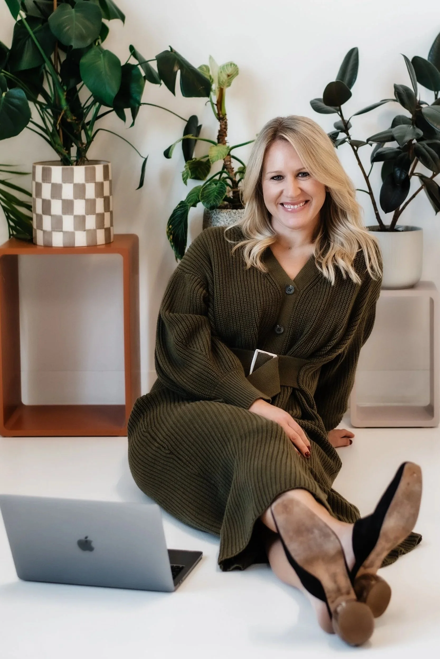 A woman with blonde hair wearing a dark green belted knit dress, sitting on the floor, smiling, with a laptop nearby, surrounded by large green houseplants in decorative pots.