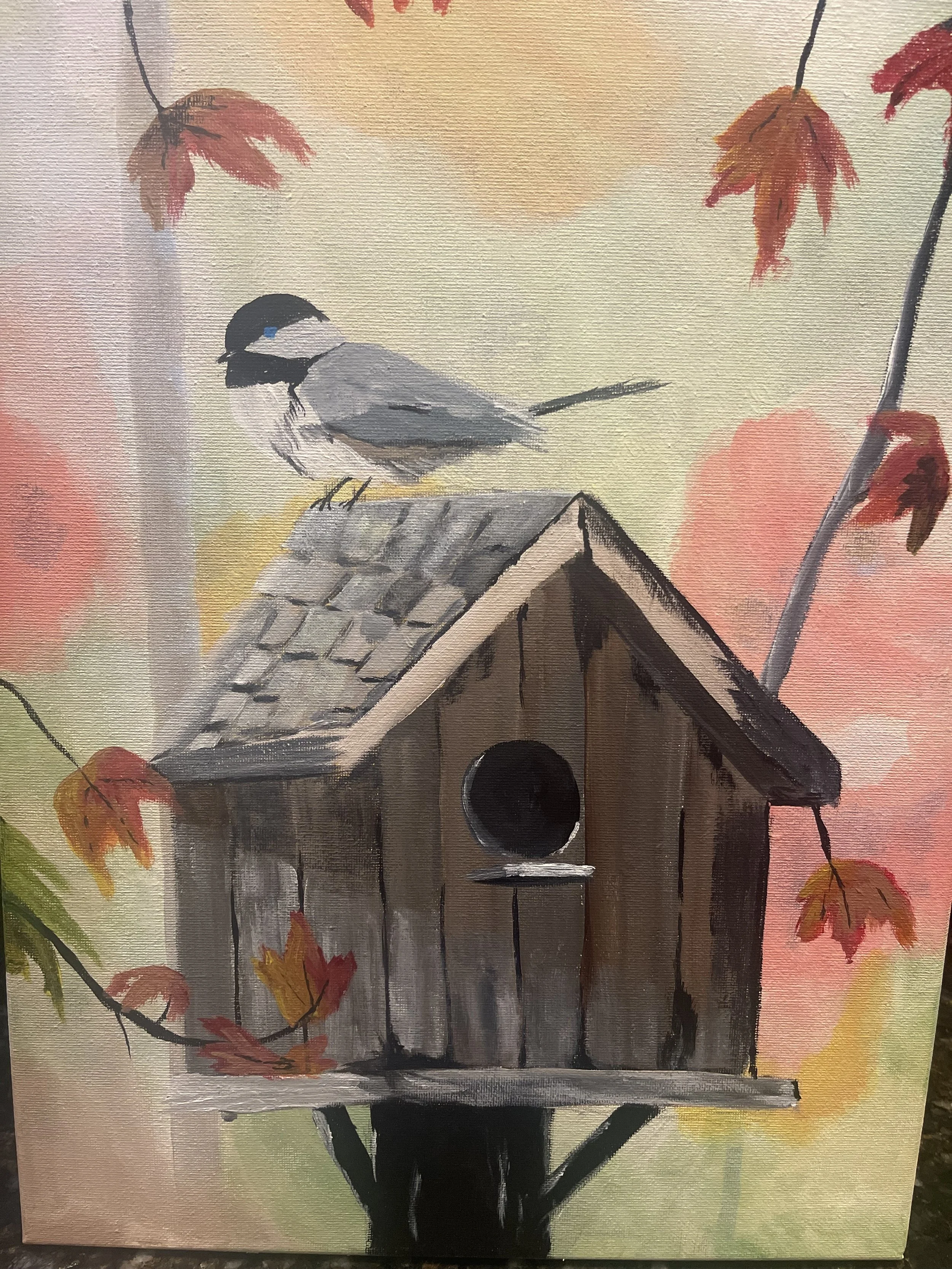 Watercolor painting of a birdhouse with a bird perched on top, surrounded by autumn leaves in shades of red, orange, and yellow.