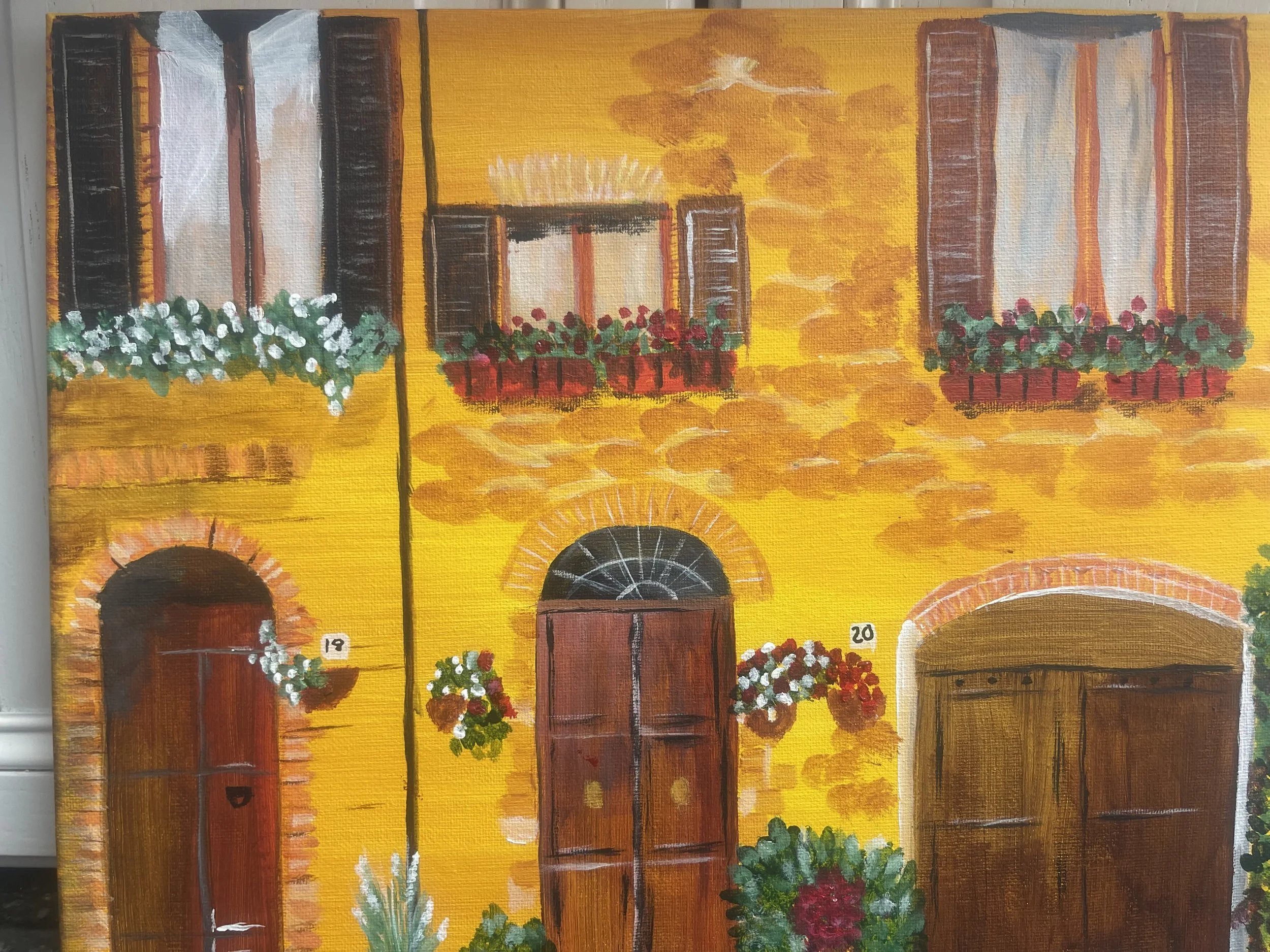 Painting of a yellow building with three brown doors numbered 19, 20, and a large arched entrance, decorated with flower boxes and green plants.
