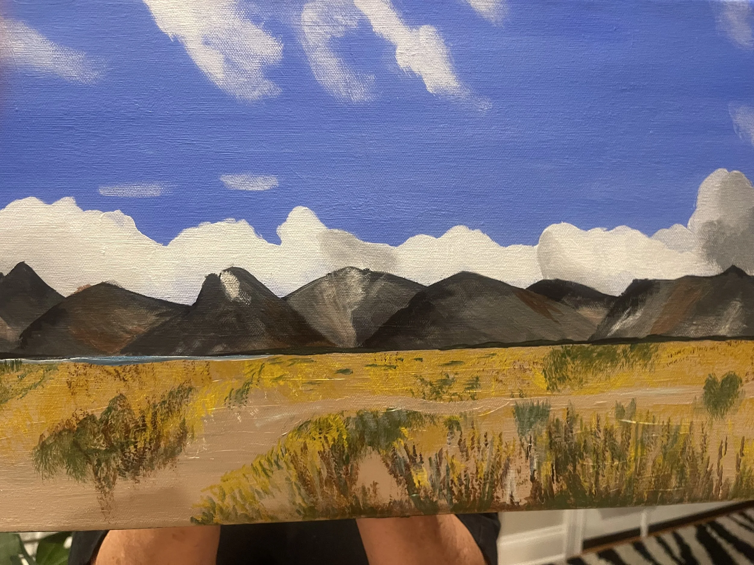 A landscape painting of mountains under a blue sky with scattered clouds, a body of water in the foreground, and shrubbery across the landscape.