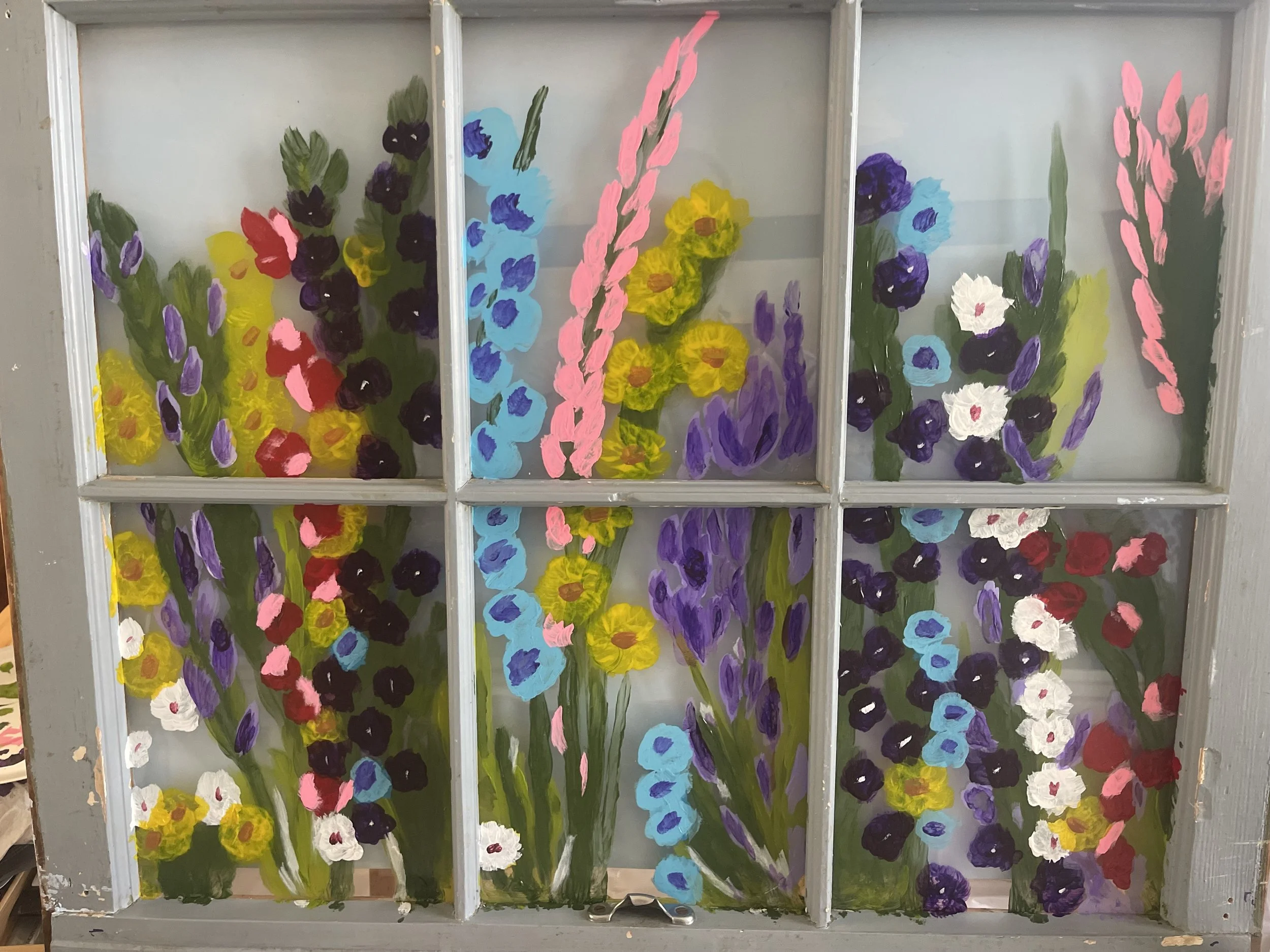 Colorful hand-painted flowers on glass panes, featuring a variety of purple, yellow, blue, red, white, and pink blossoms with green stems and leaves, framed by a wooden window structure.