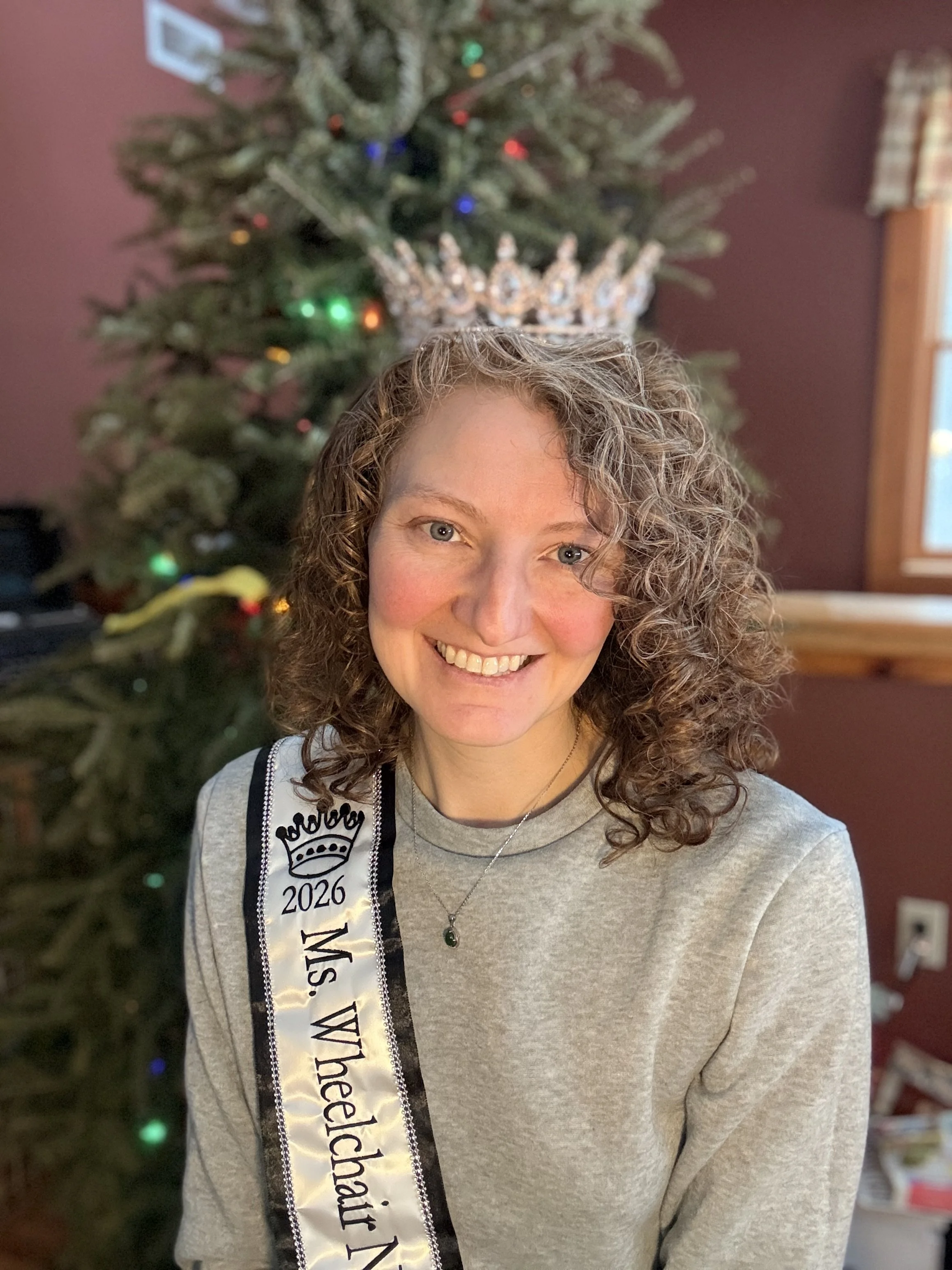 A head shot of Nenia, a white woman with red curly hair. She is wearing a silver crown and a sash that reads 2026 Ms. Wheelchair NH.