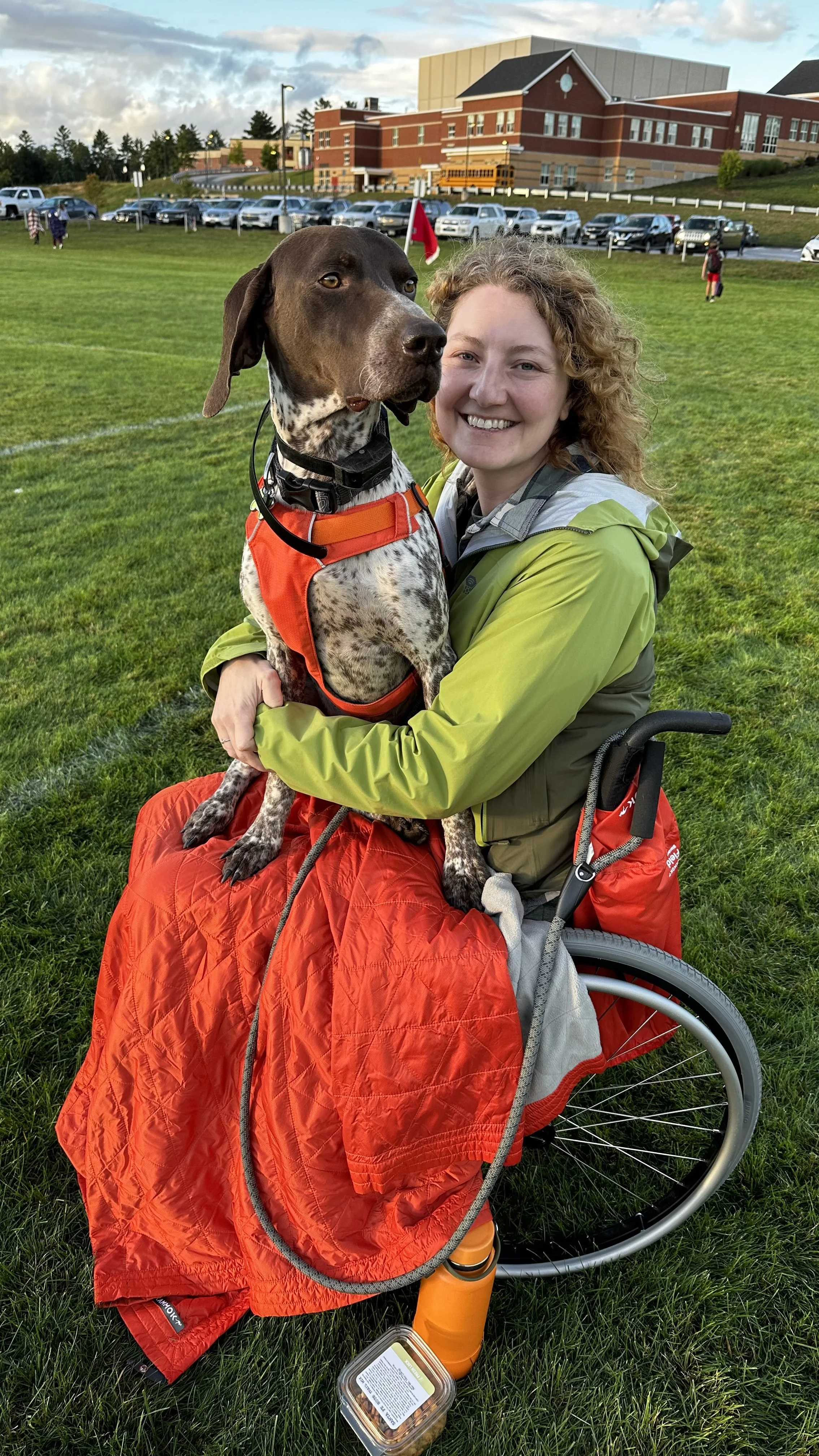 Nenia, a white woman in a manual wheelchair, sits smiling at the camera, holding her brown and white German Short Hair Pointer on her lap.