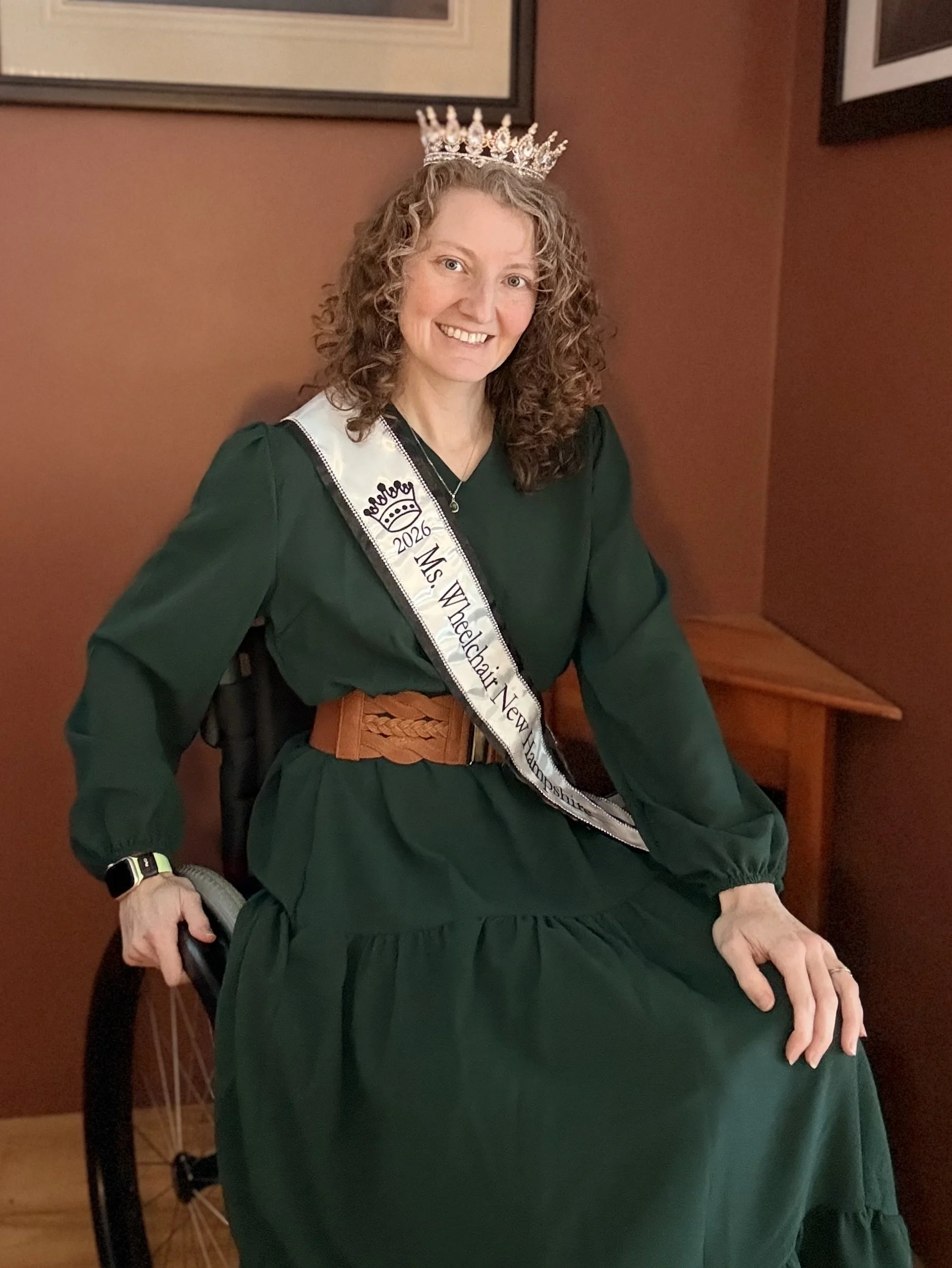 Nenia, a white woman in a manual wheelchair, wearing a long green dress, smiling at the camera with a sash that says Ms. Wheelchair New Hampshire 2026 and wearing a silver crown on her head. She has curly red hair.