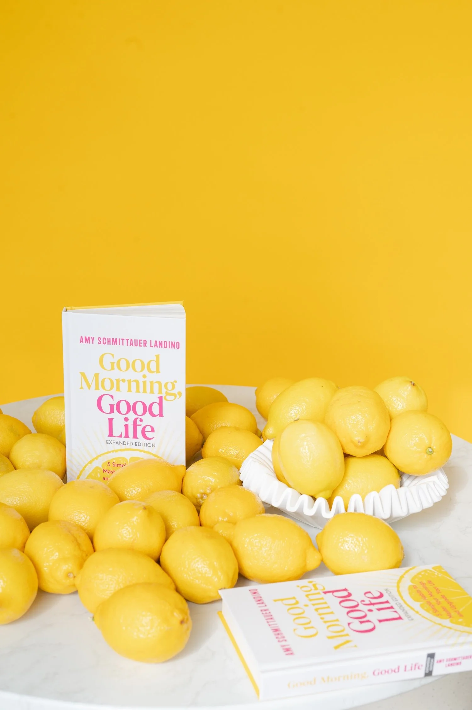 A product shot of Amy Schmittauer Landino's book Good Morning, Good Life. The book is standing on a table surrounded by lemons set on a yellow backdrop.