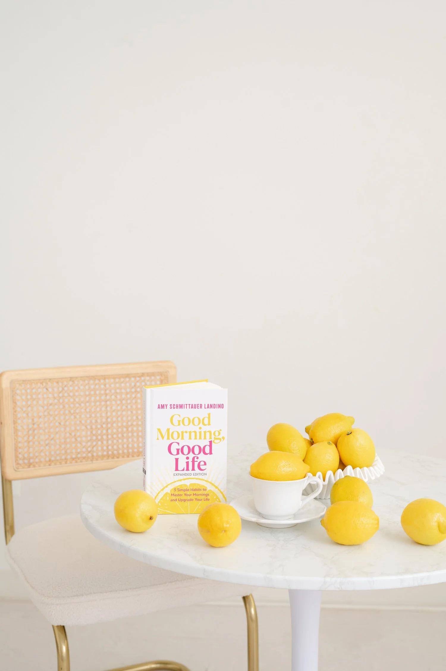 A book titled Good Morning, Good Life by Amy Schmittauer Landino is standing up on a white table, with yellow lemons surrounding it. The scene is set on an off-white background.