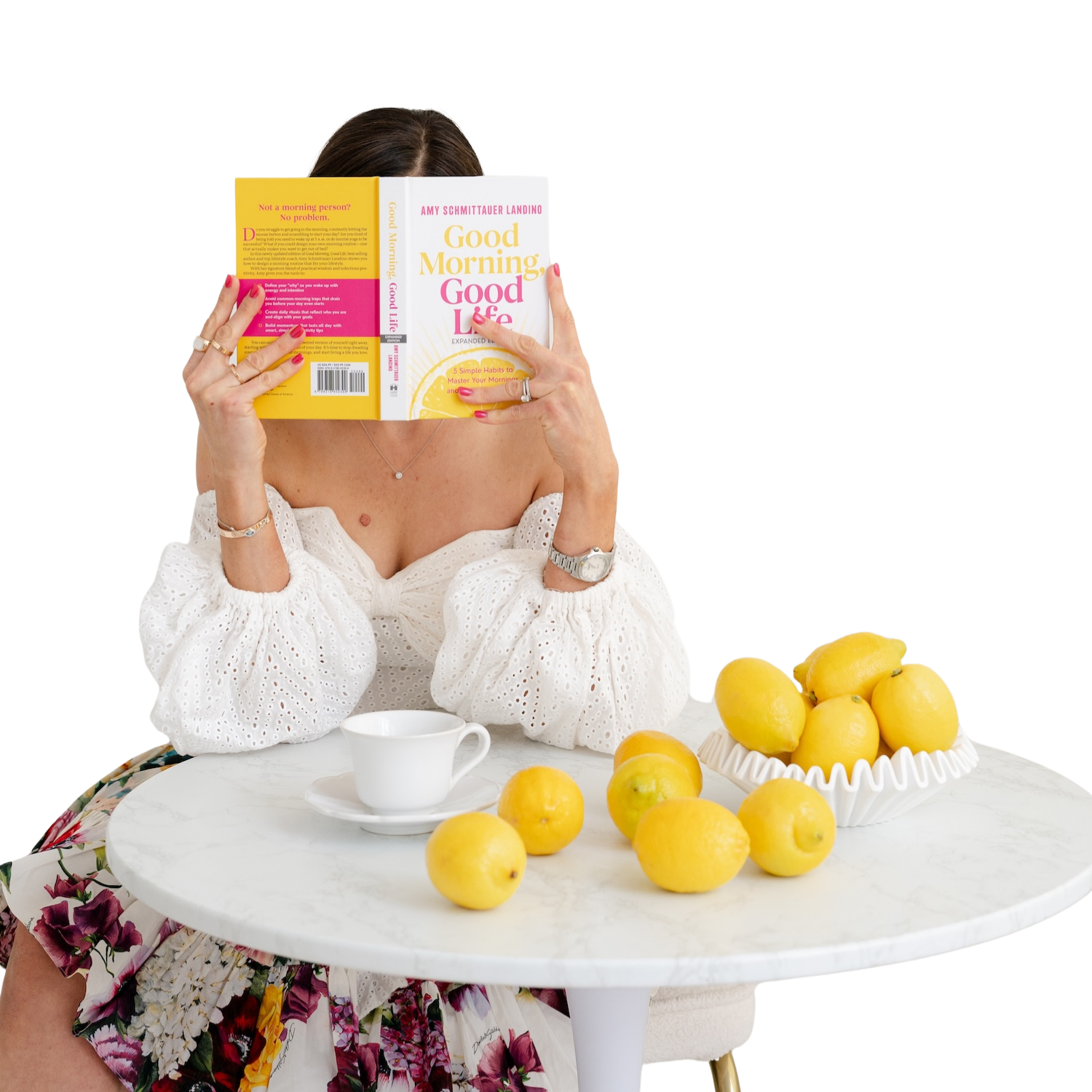 Author Amy Schmittauer Landino, sits at a table covered with lemons. She's wearing a white blouse and holding up her book, Good Morning, Good Life in front of her face.