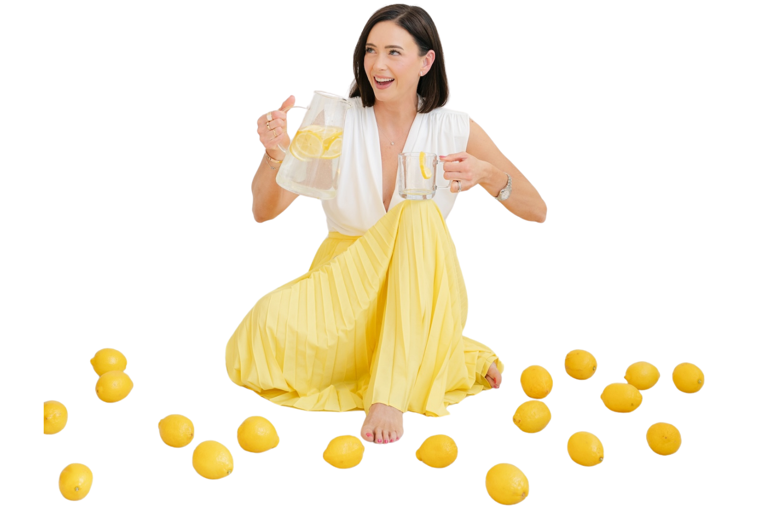 Amy Landino, author of Good Morning, Good Life, is sitting among a pile of lemons as she pours a glass of lemon water from a glass pitcher.