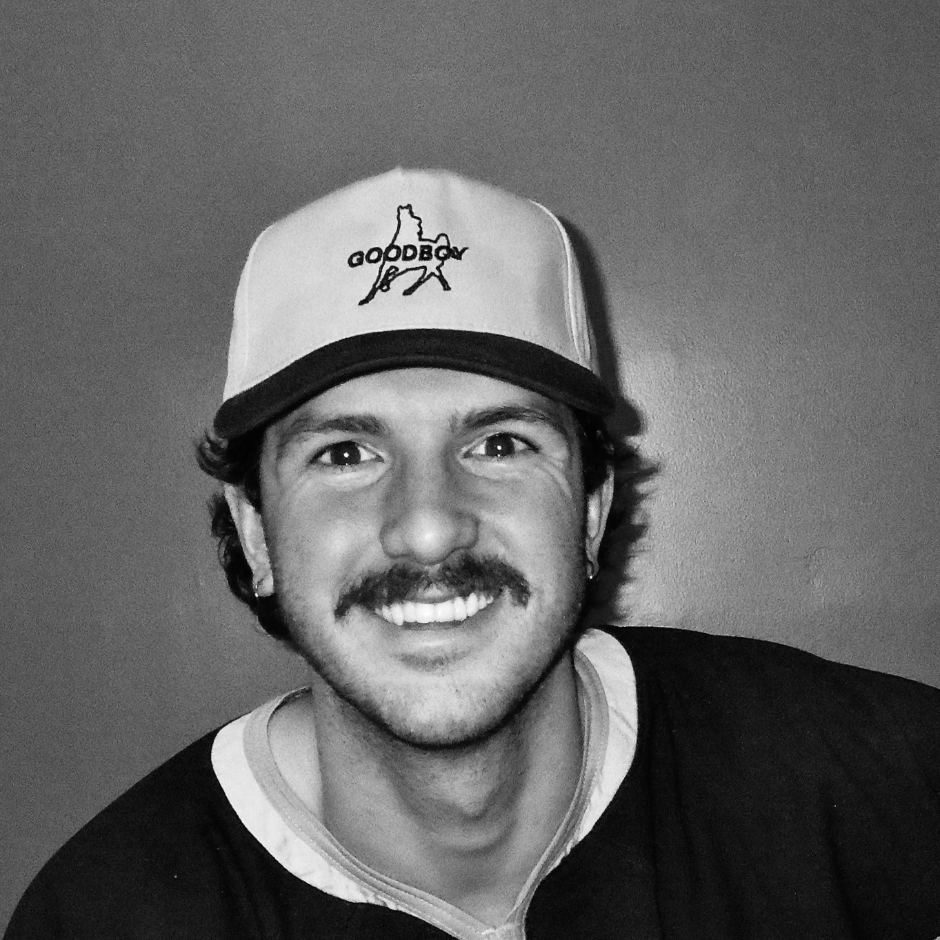 A smiling man wearing a baseball cap with the word 'Goodbar' and a mountain outline on it, a black shirt, and earrings, against a plain background.