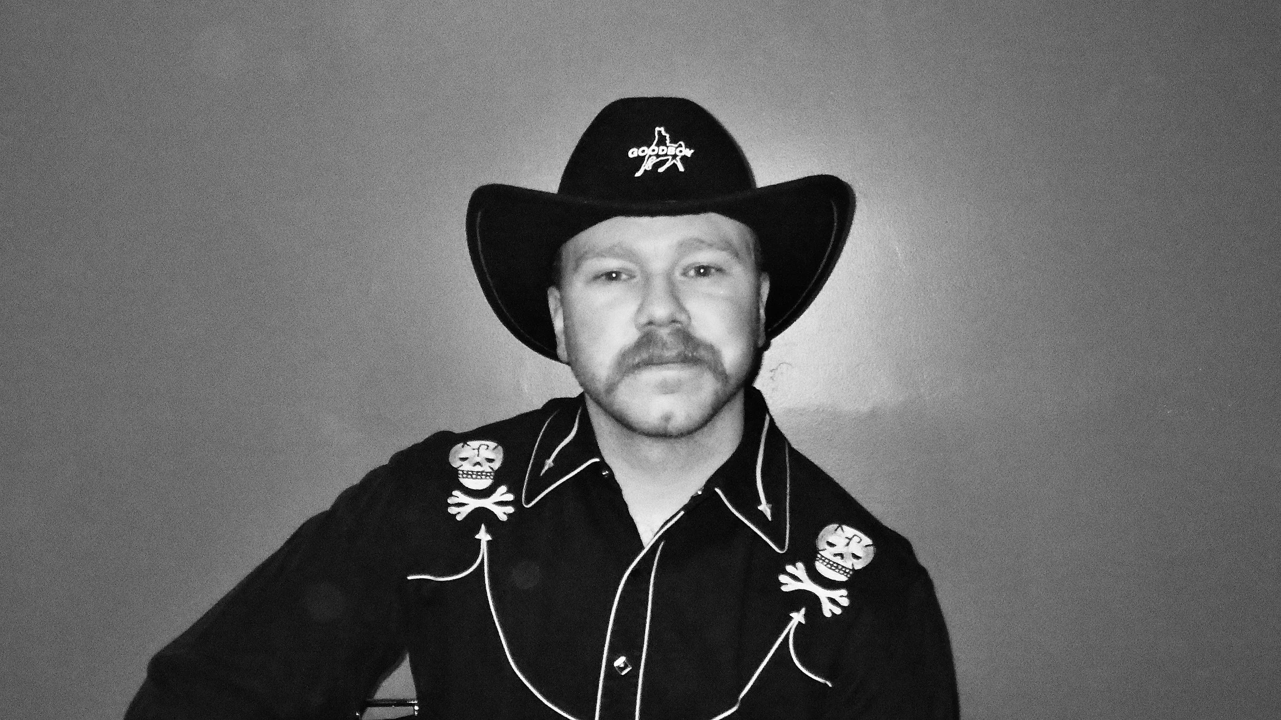 A man wearing a cowboy hat and a western-style shirt with skull and crossbone patches, posing against a plain wall.