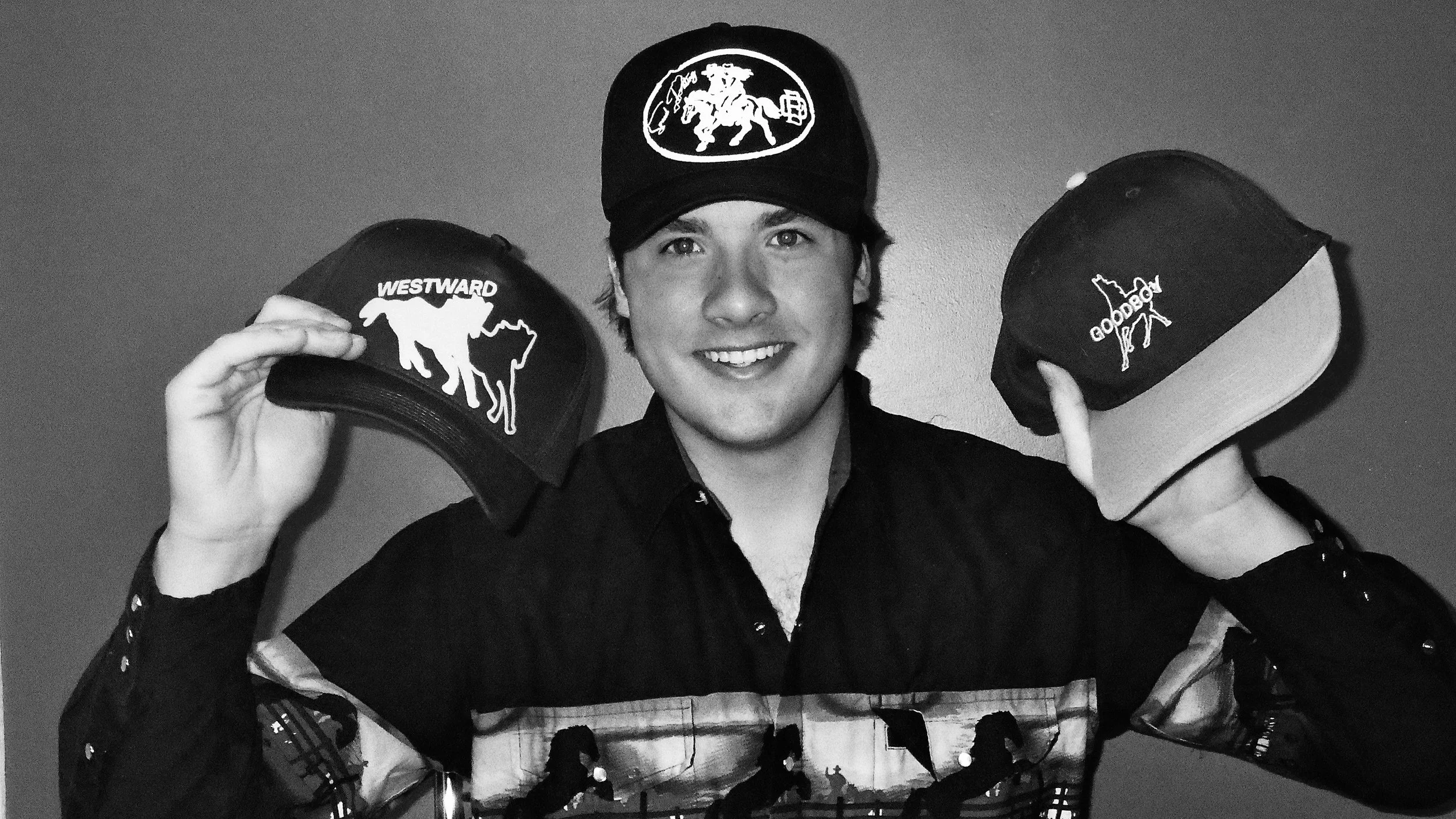 A young man smiling, wearing a cap and holding up three caps with different logos and designs.