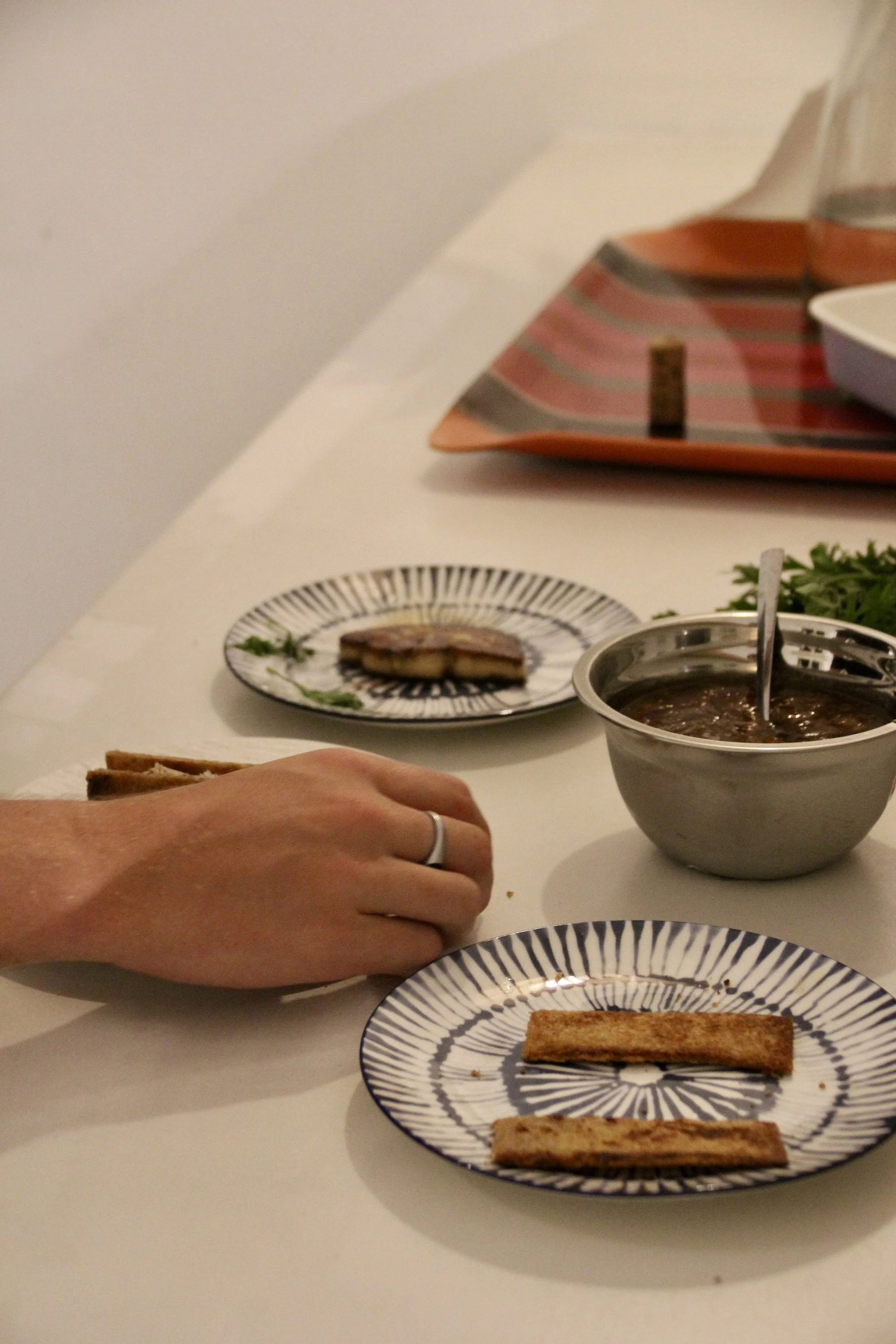 Une main posant une tranche de pain grillé sur une petite assiette à rayures bleues et blanches, avec une soupe dans un bol en acier inoxydable à côté et une autre assiette avec une tranche de viande ou de poisson en arrière-plan.