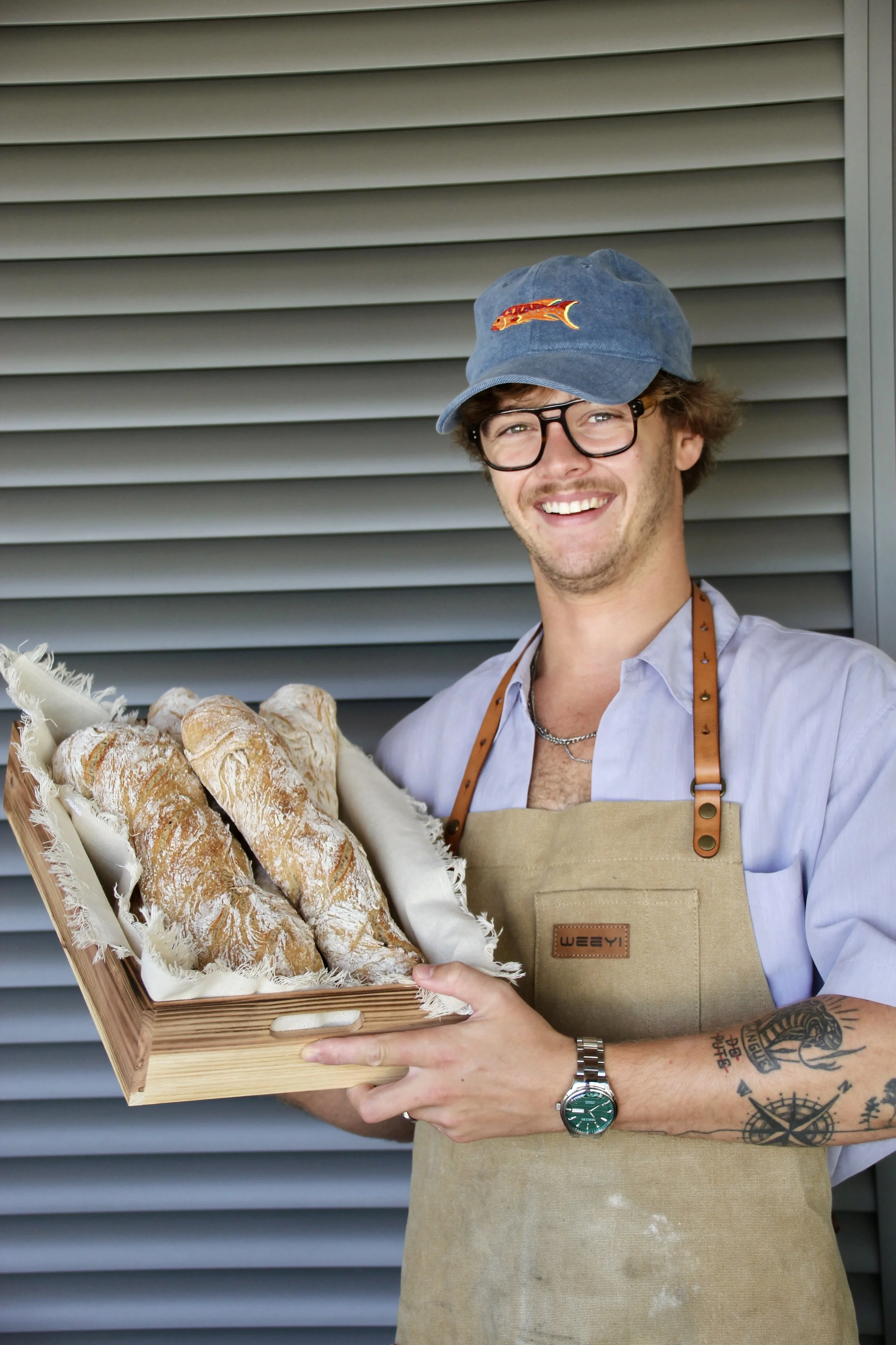 Jeune homme souriant portant une casquette, des lunettes et un tablier dans la main un panier de pains baguettes