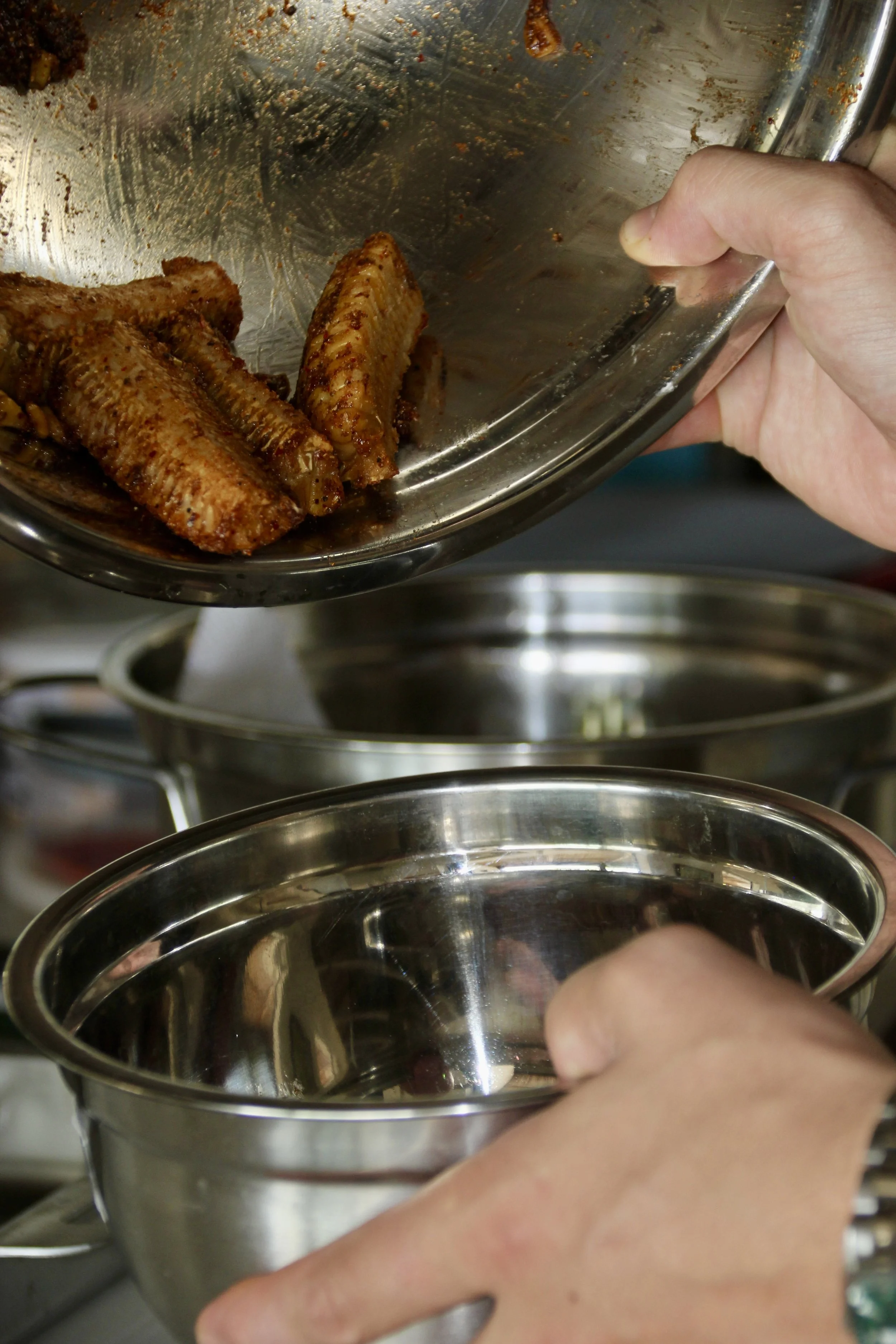Deux mains tiennent des bols en acier inoxydable, portant des morceaux de viande assaisonnée. La scène se déroule en cuisine.