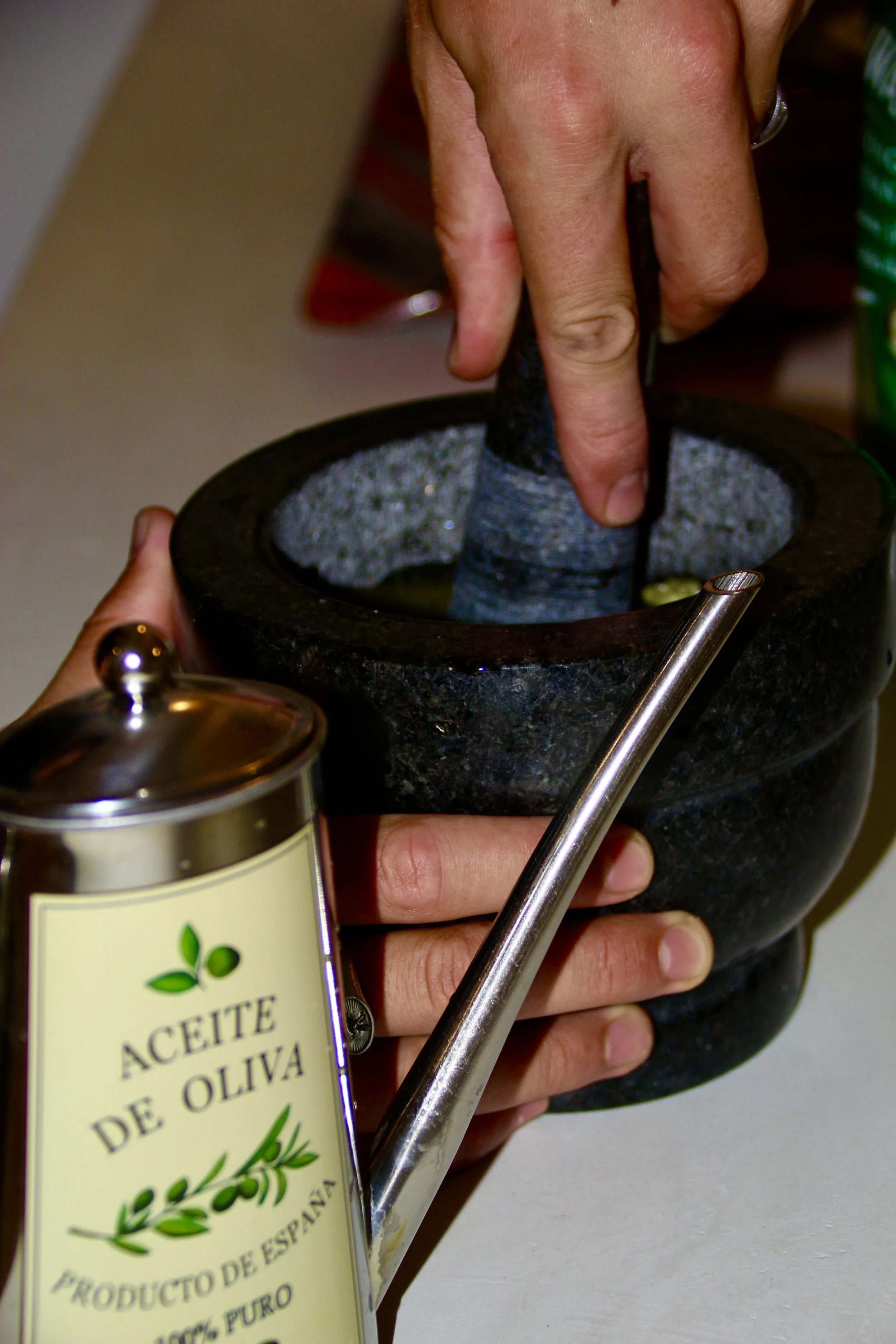 Une main tenant un mortier en pierre et une autre main utilisant un pilon pour moudre des ingrédients, avec une bouteille d'huile d'olive à proximité.