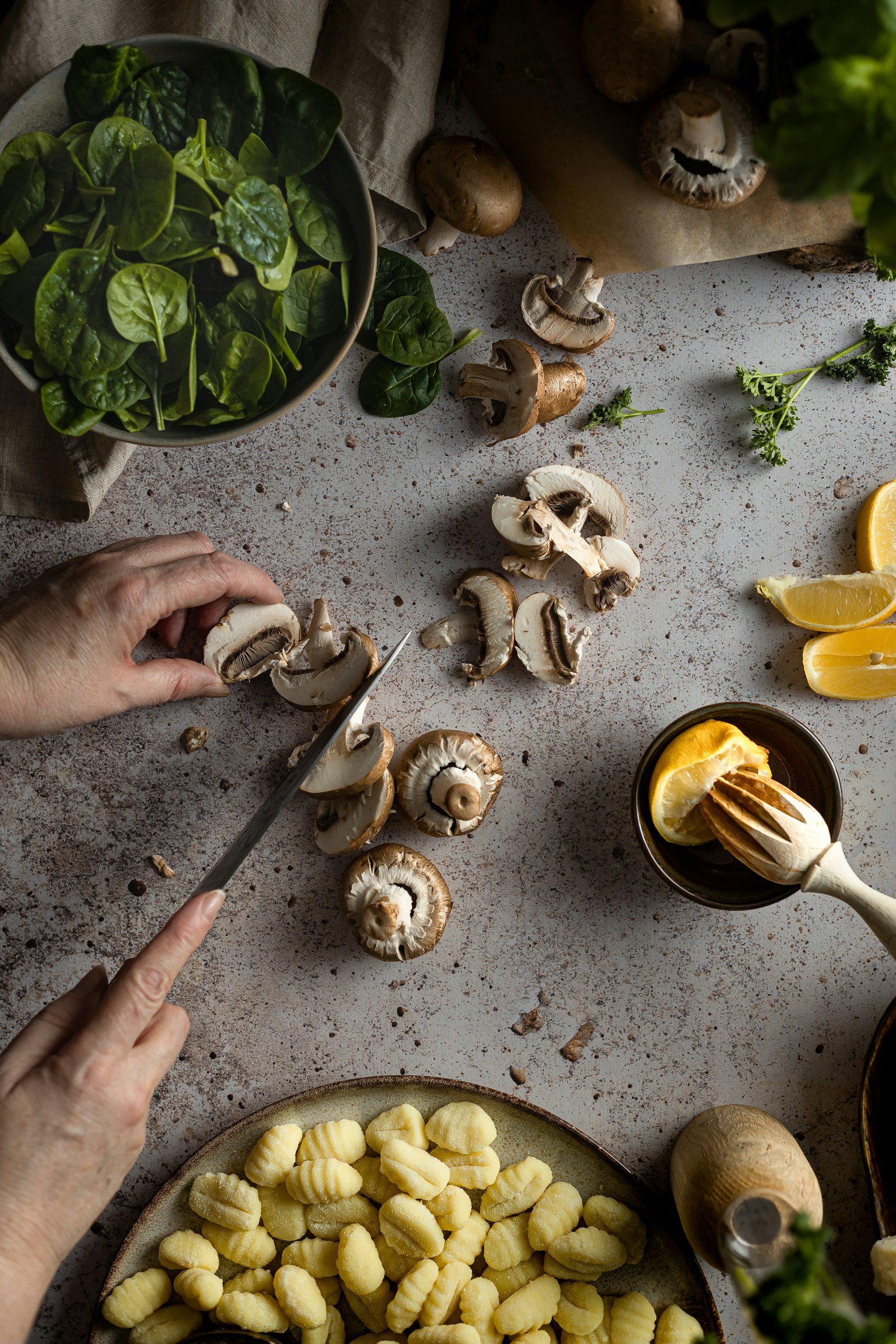 Une personne coupe des champignons dans une cuisine, entourée de légumes verts, de citrons, de gnocchis, et d'autres ingrédients pour cuisiner.