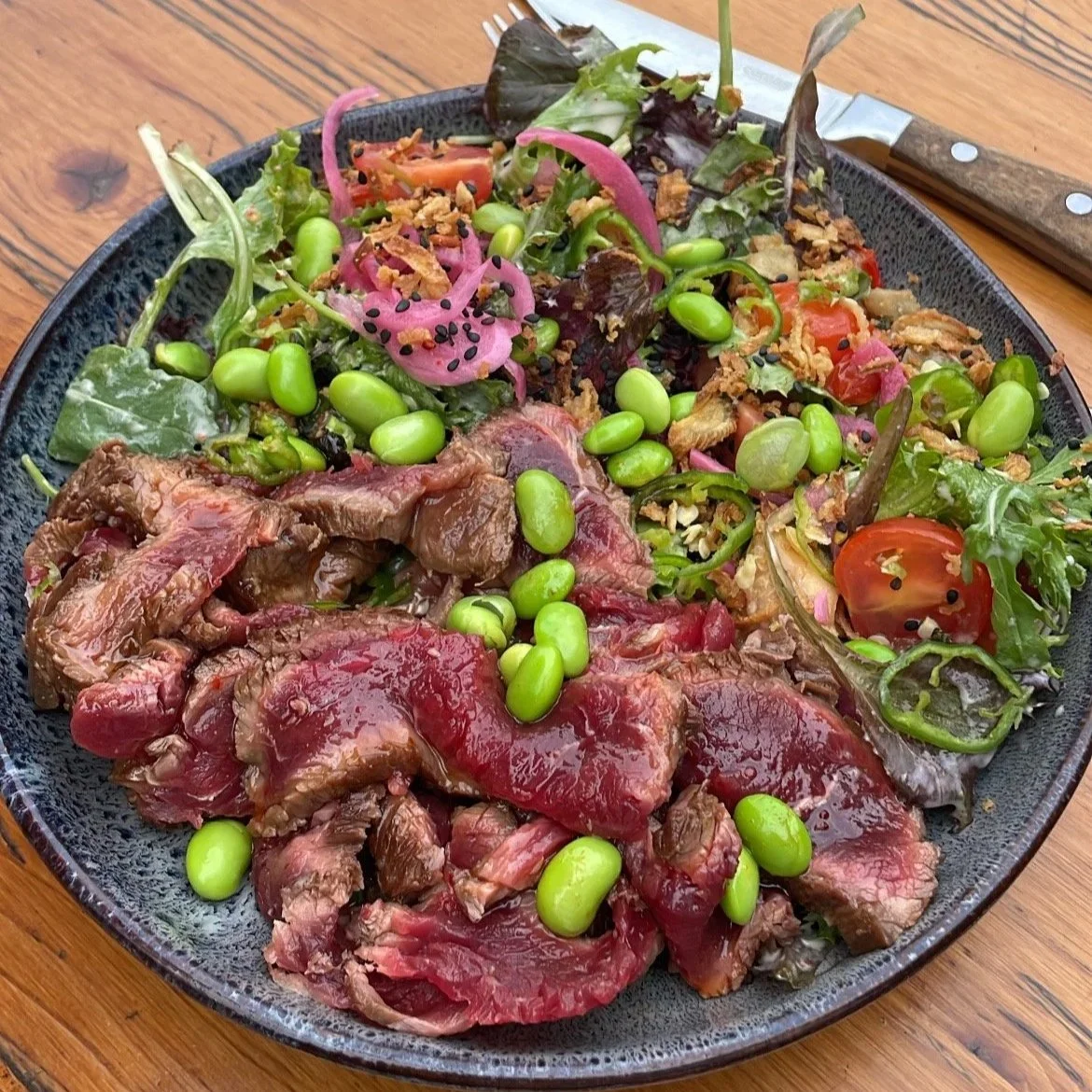 Salade composée avec de la viande, des tomates, des edamames, des oignons roses, et d'autres légumes, dans un bol noir sur une table en bois.