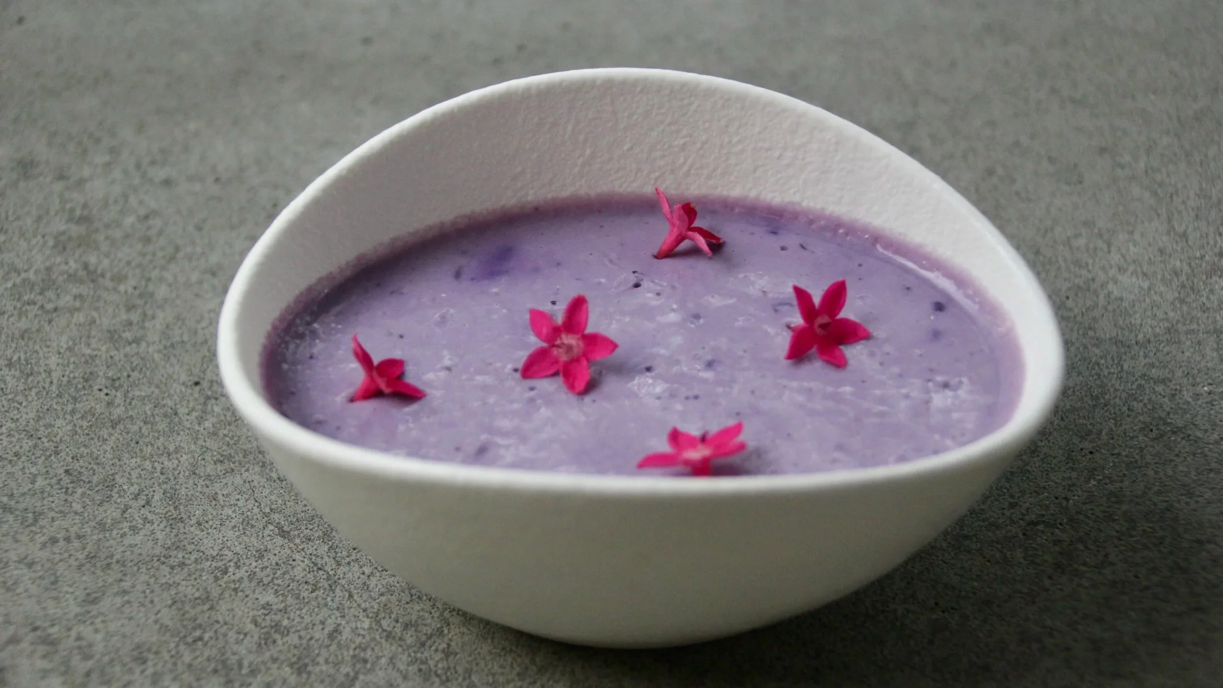 Soupe violette garnie de petites fleurs roses dans un bol blanc sur une surface grise.