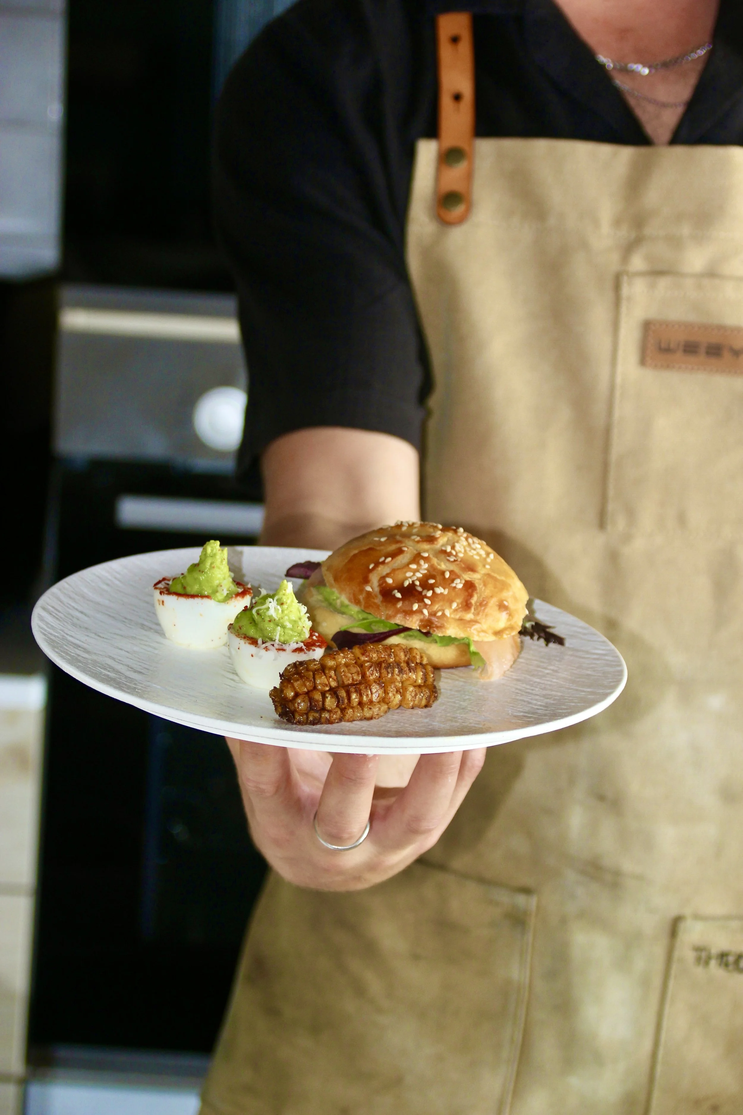 Personne tenant une assiette avec un sandwich, des œufs farcis à l'avocat et un épi de maïs grillé.