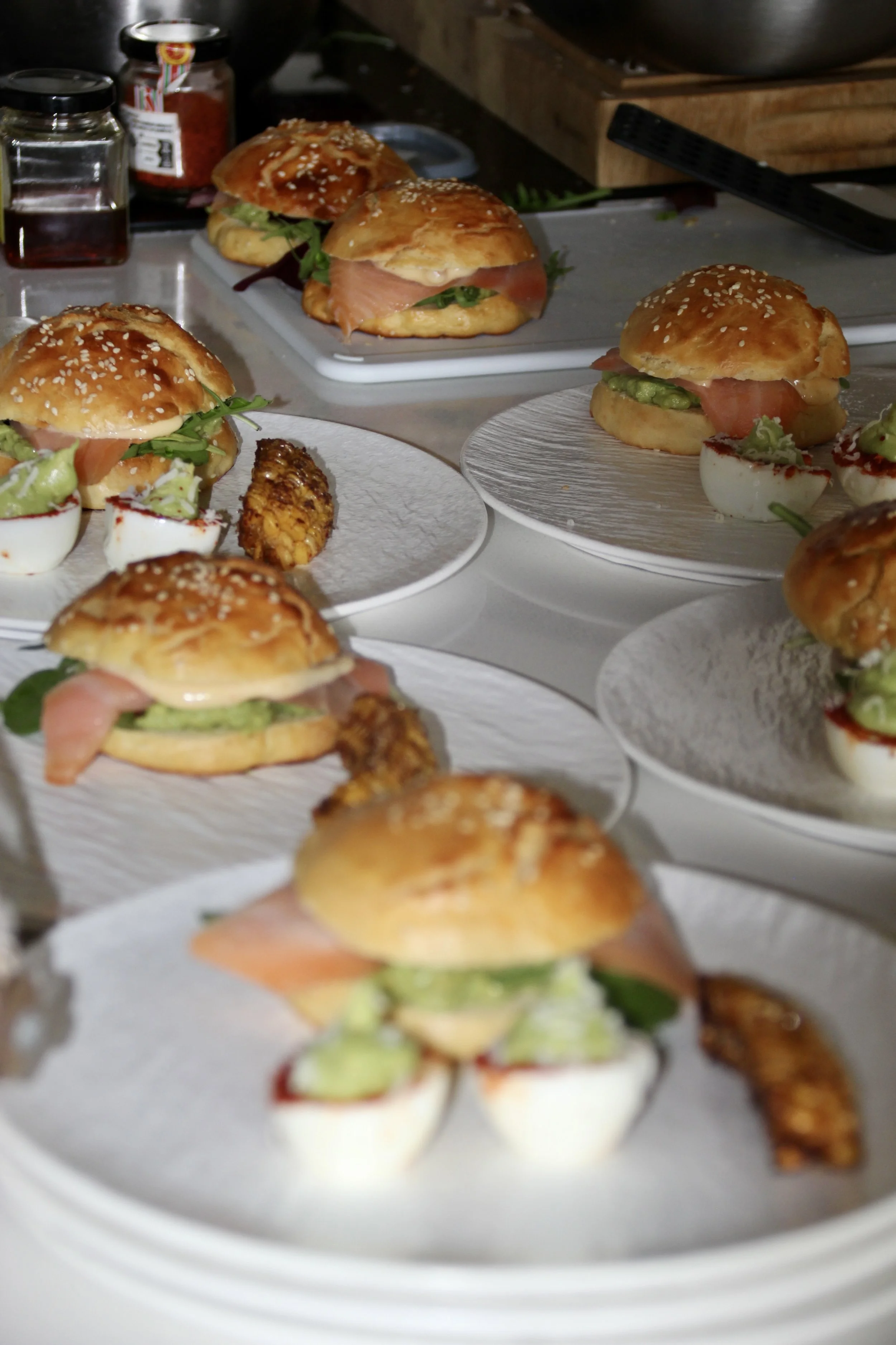 Mini sandwich burgers with smoked salmon, lettuce, and avocado, served on white plates with sesame seed buns.