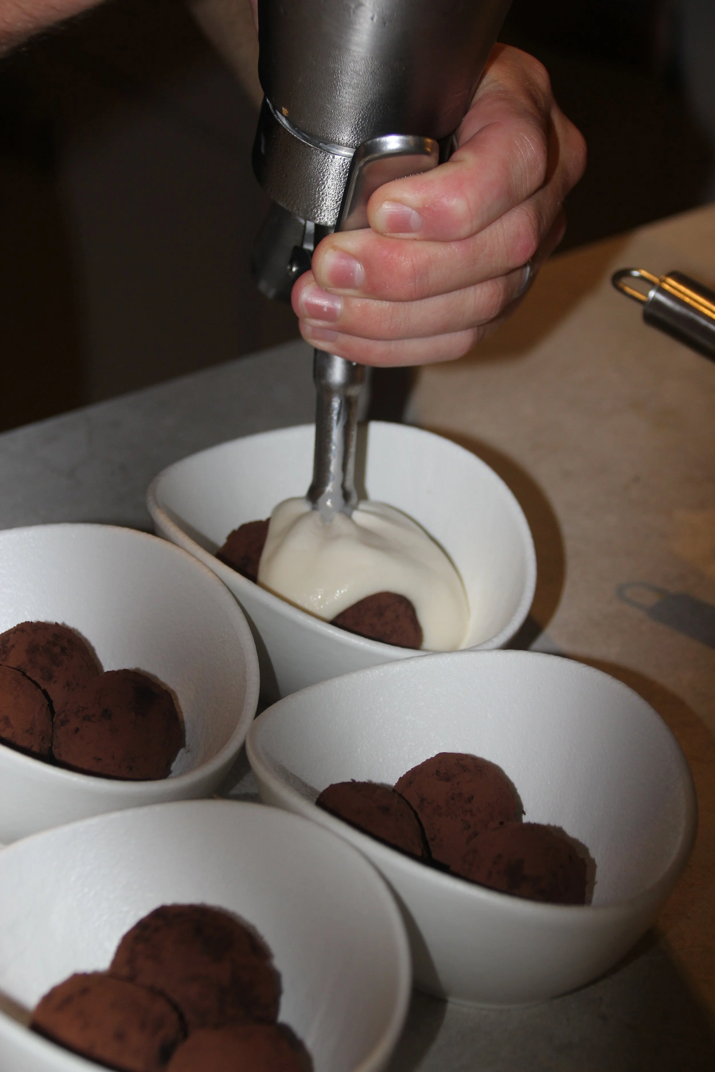 Une personne utilisant un siphon pour ajouter de la crème sur des truffes au chocolat dans des bols blancs.