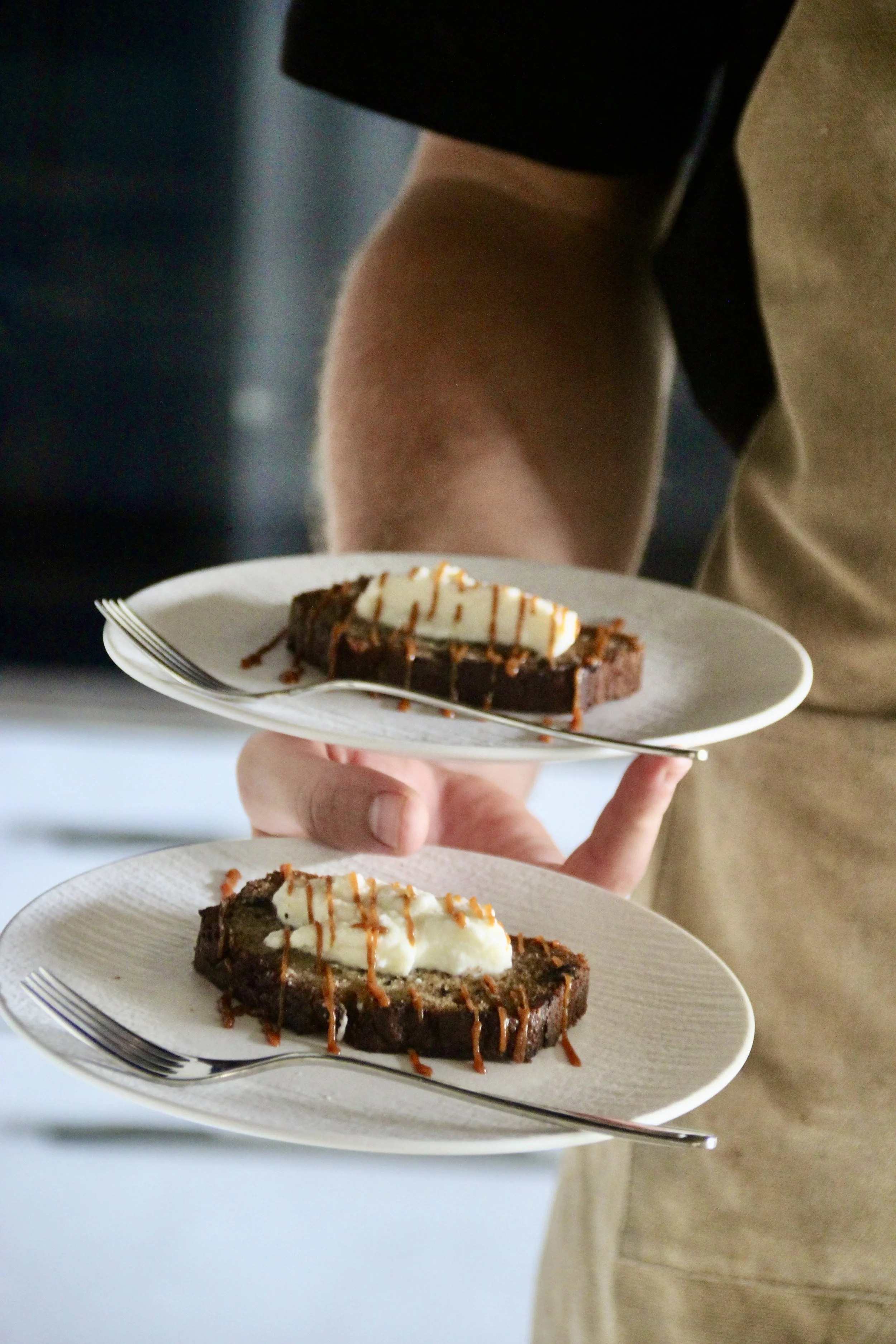 Deux assiettes de gâteau à la crème, garnies de caramel, tenues par une personne portant un t-shirt noir et un bermuda beige.