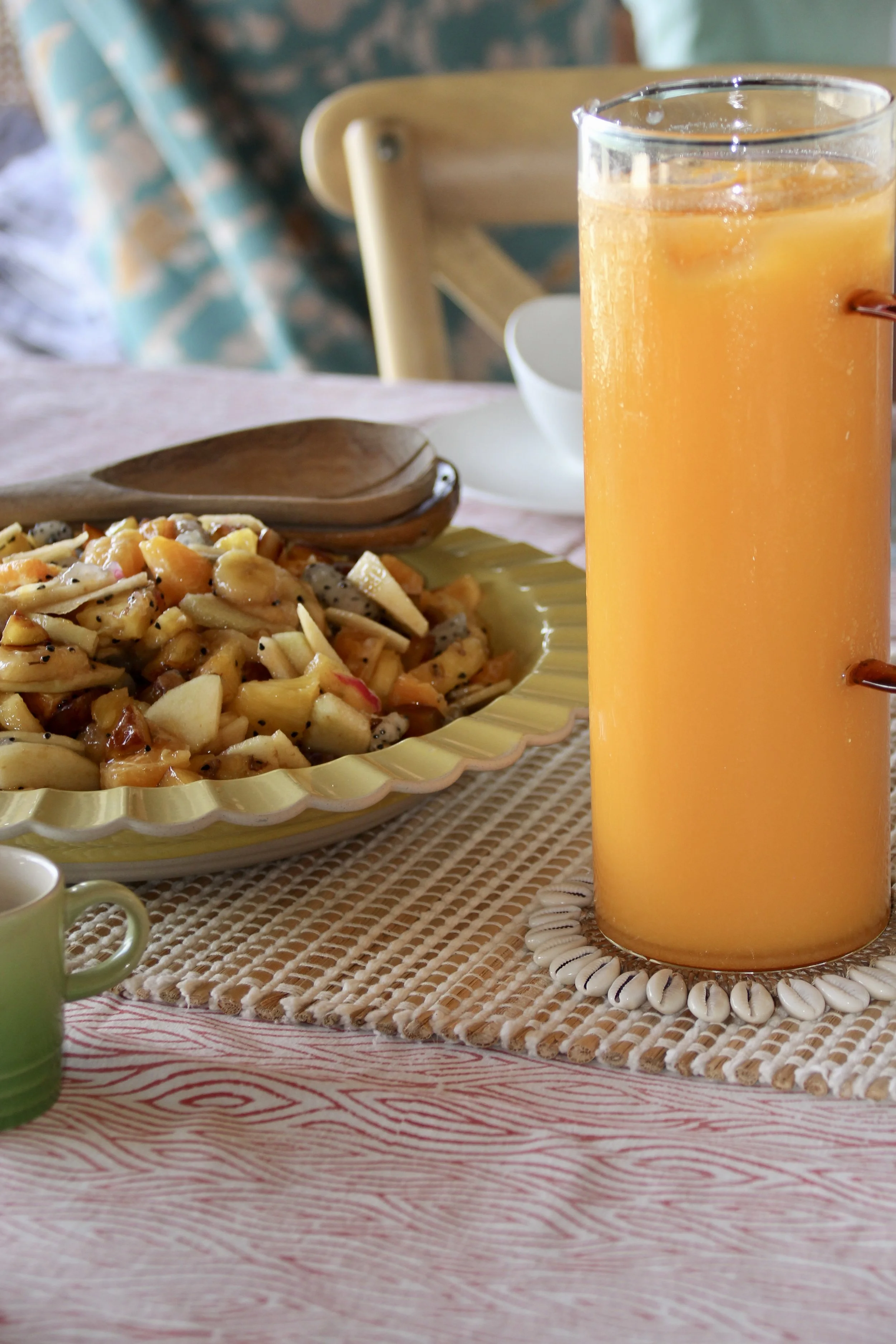 Verre d'orange pressée, assiette de fruits frais coupés, tasse blanche, nappe rose, assiette jaune, cuillère en bois, décoration de plage avec coquillages