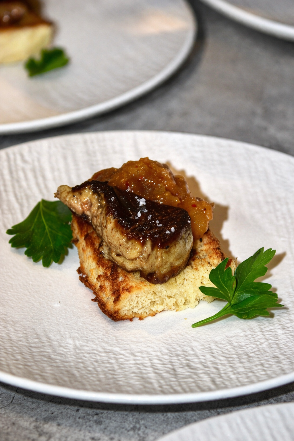 Une assiette avec un morceau de pain grillé, une pièce de foie gras, une compote de figues, garnie de persil et de sel.