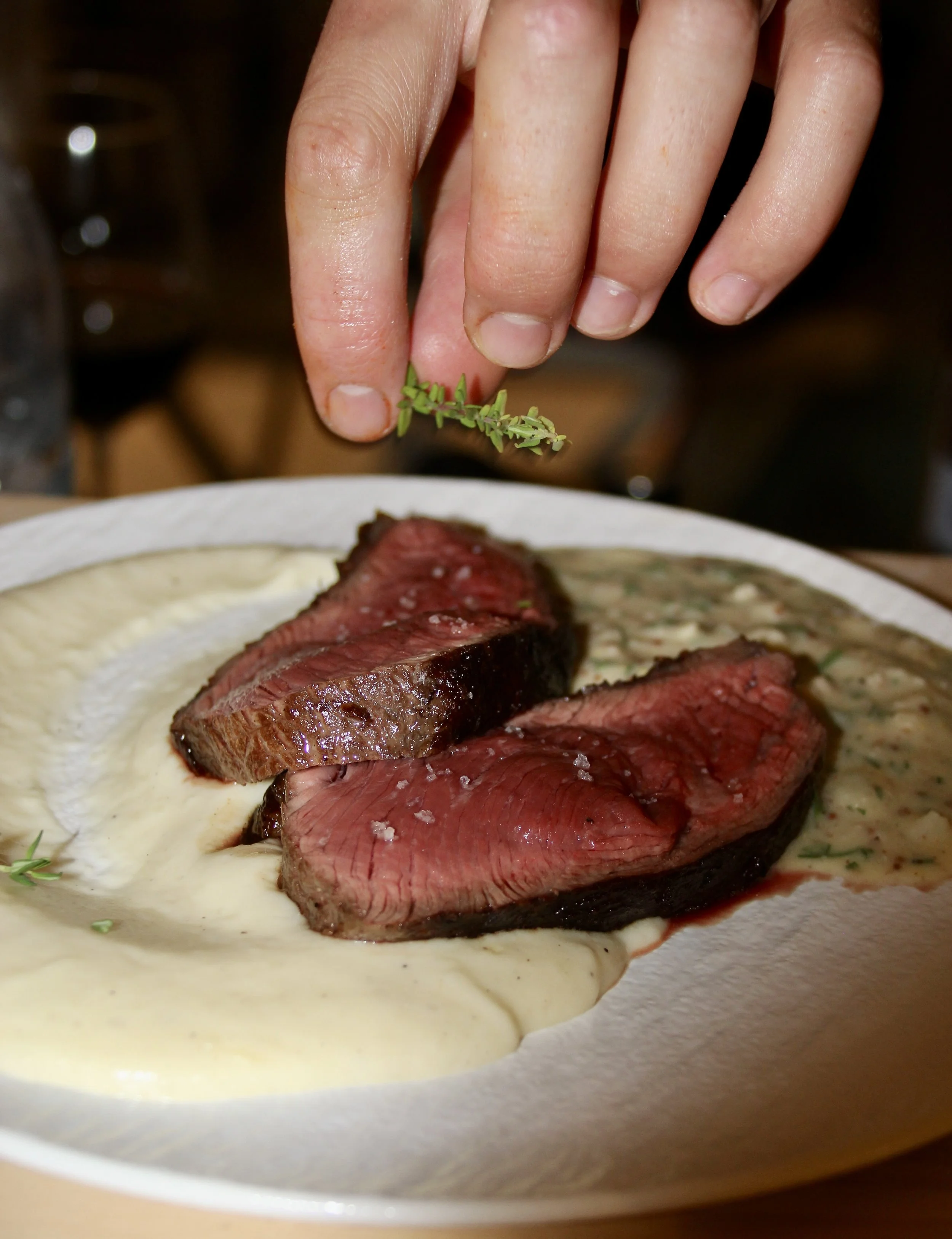 Une main saupoudre de fines herbes sur un plat de viande saignante accompagnée de purée et de sauce.