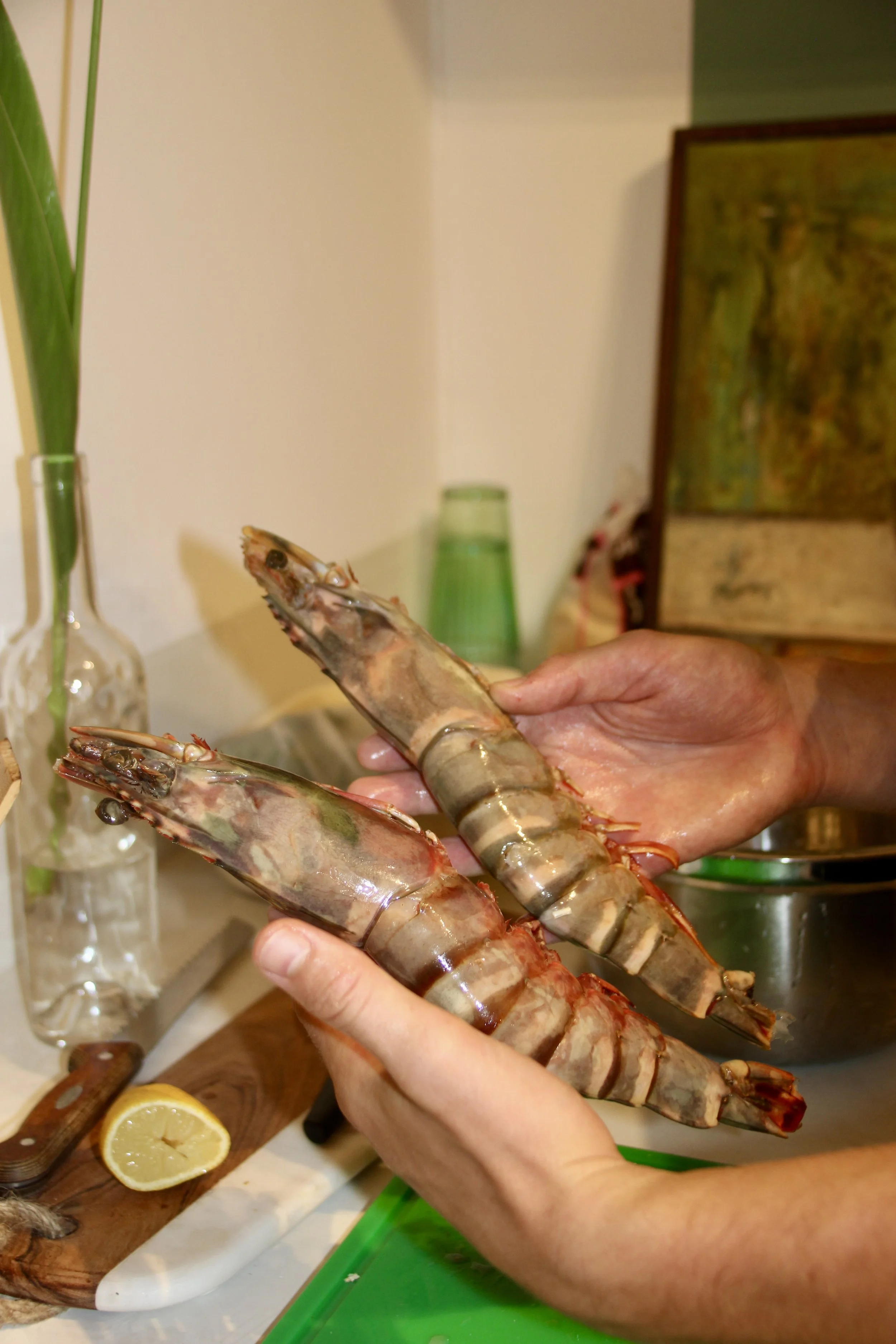 Des mains tiennent deux grosses crevettes crues au dessus d'un plan de travail de cuisine. Un demi citron et un couteau en bois sont visibles à côté.