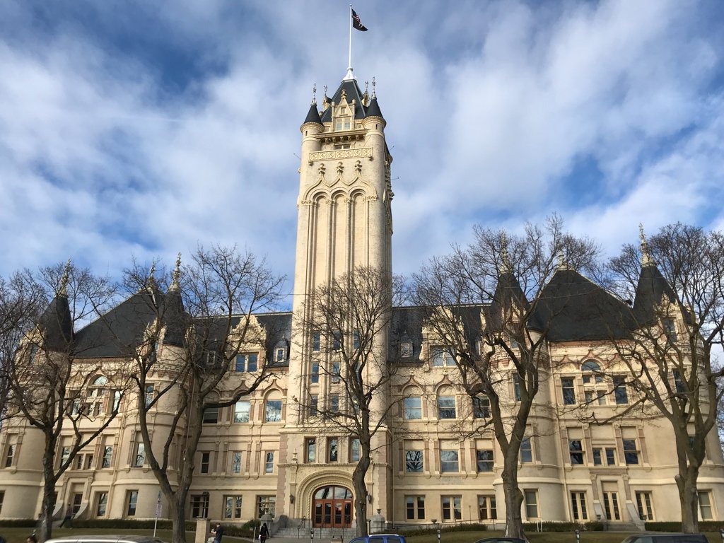 A large castle-like historic building with a tall central tower topped by a flag, surrounded by leafless trees and a partly cloudy sky in the background.