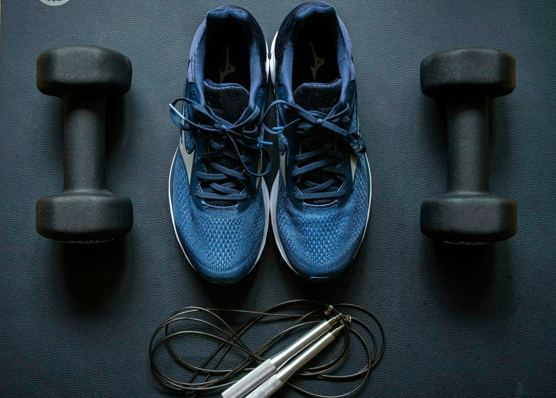 A pair of blue running shoes, two black dumbbells, and a jump rope with silver handles on a black textured exercise mat.