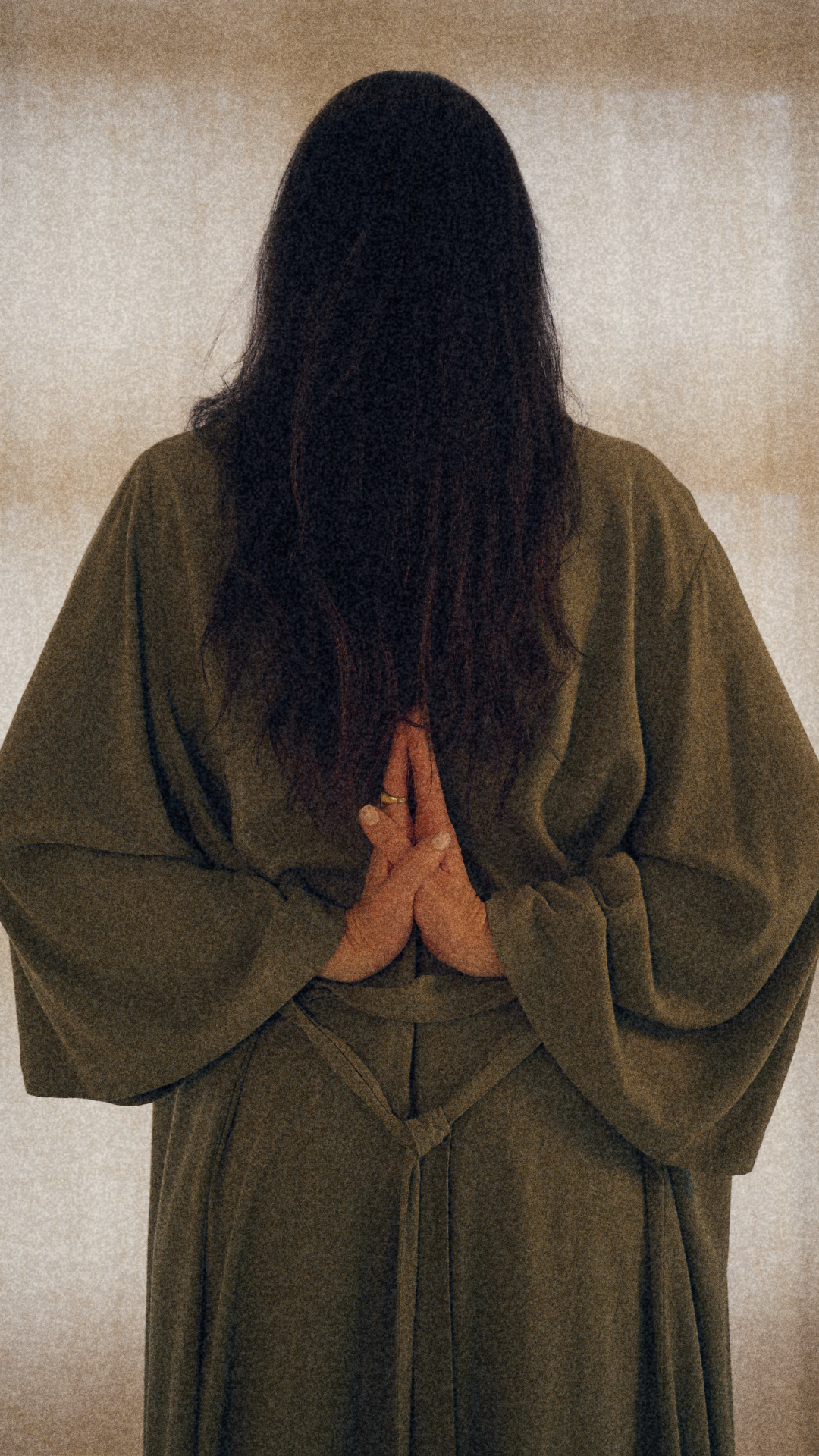 A person with long dark hair, wearing loose-fitting green clothing, standing against a neutral background, with hands pressed together in a prayer or greeting gesture.