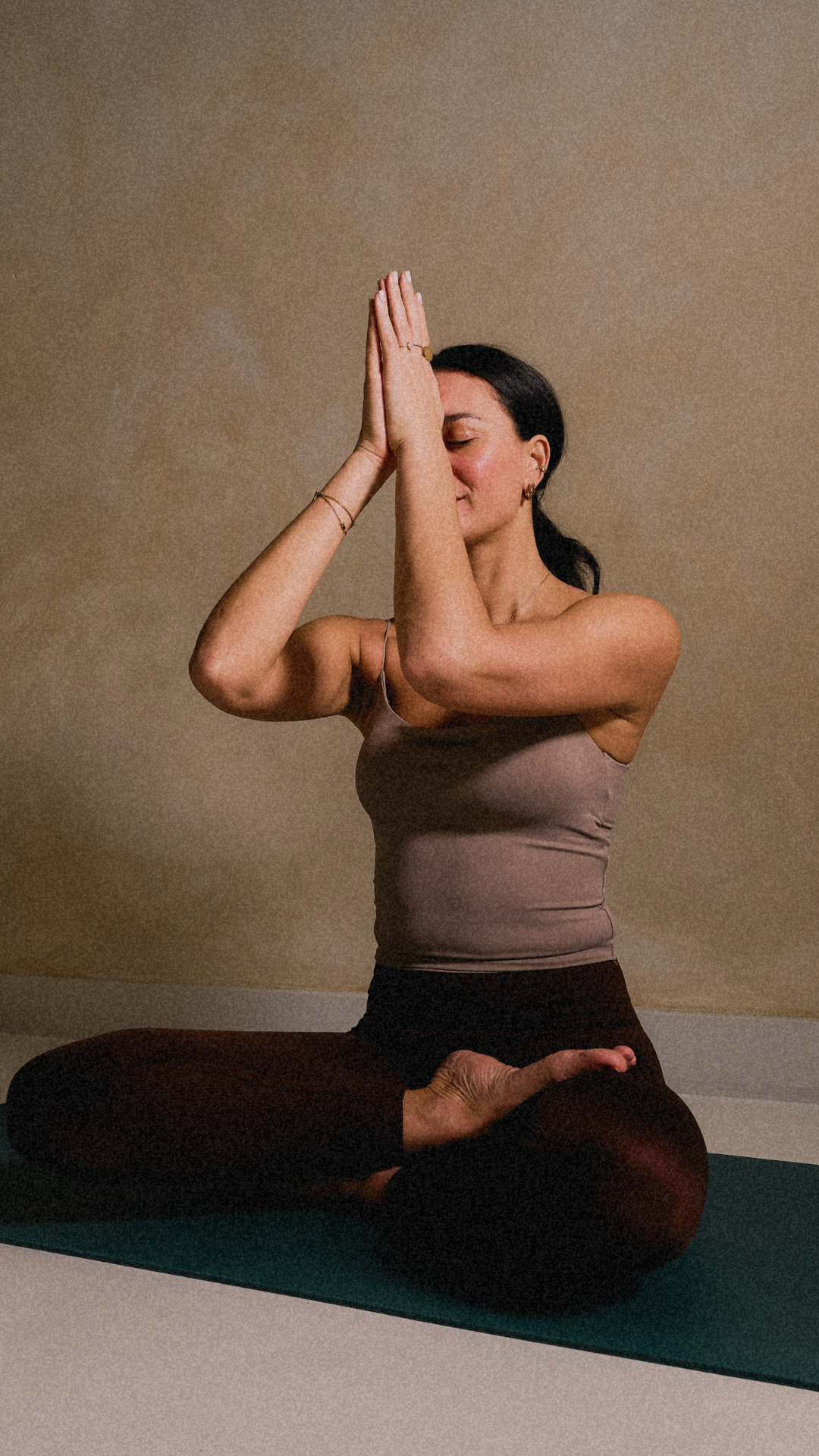 A woman practicing yoga in a seated meditation pose with palms pressed together in front of her face, eyes closed, on a green yoga mat against a plain beige wall.