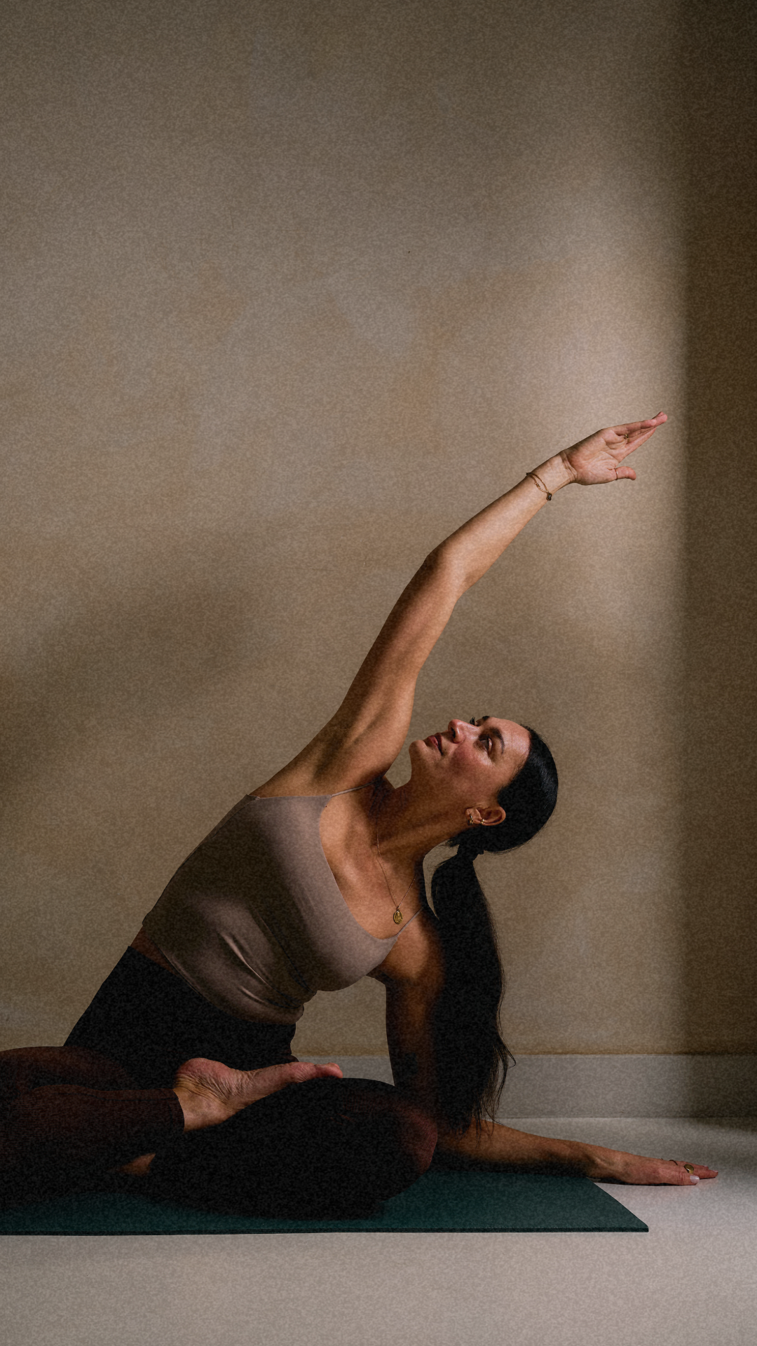 A woman practicing yoga indoors, seated on a mat, reaching upward with her right arm while looking in that direction, with a beige wall backdrop.