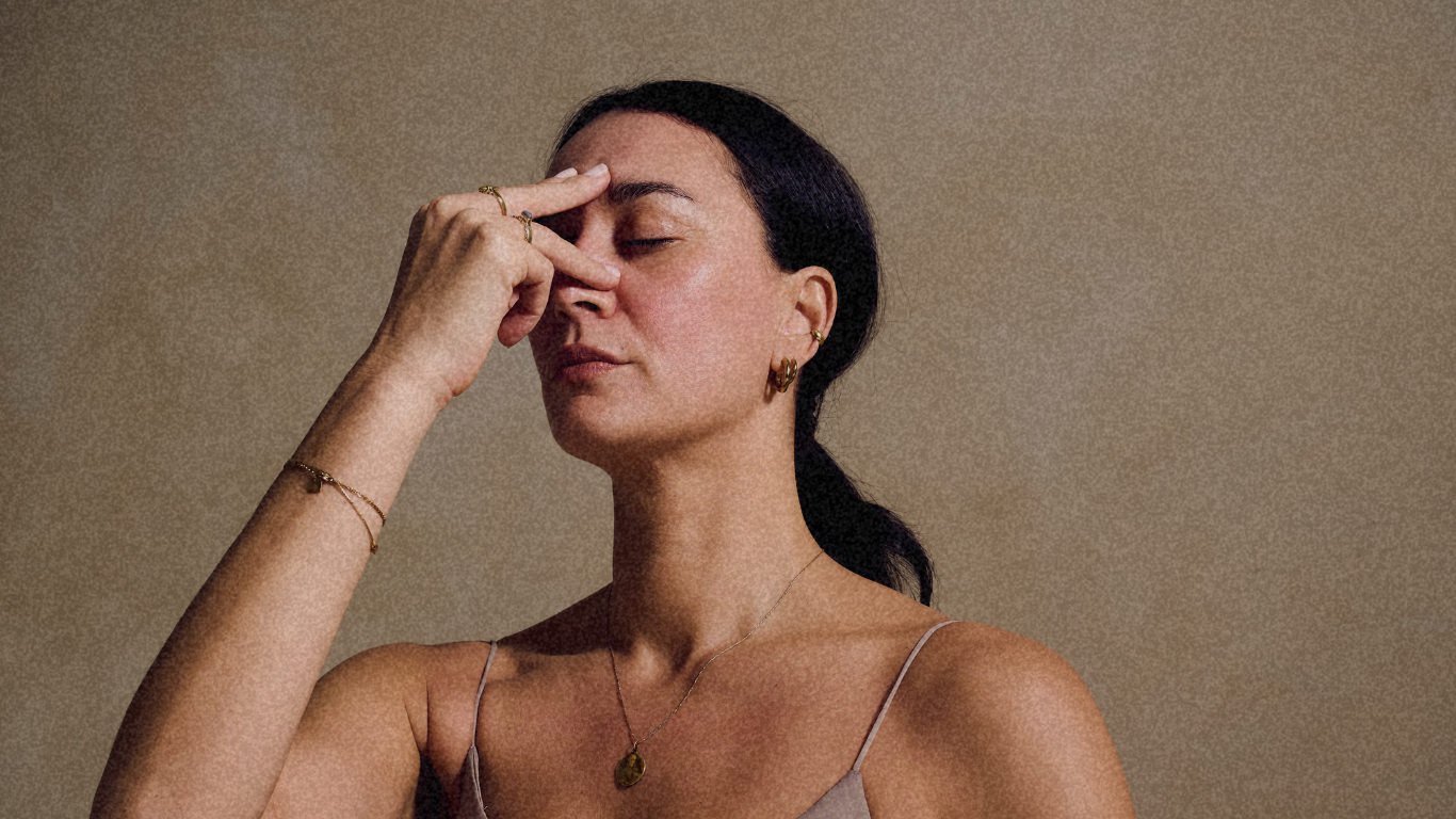 A woman with black hair, wearing a beige camisole, gold jewelry, and her eyes closed, holding her hand to her forehead in a contemplative pose.