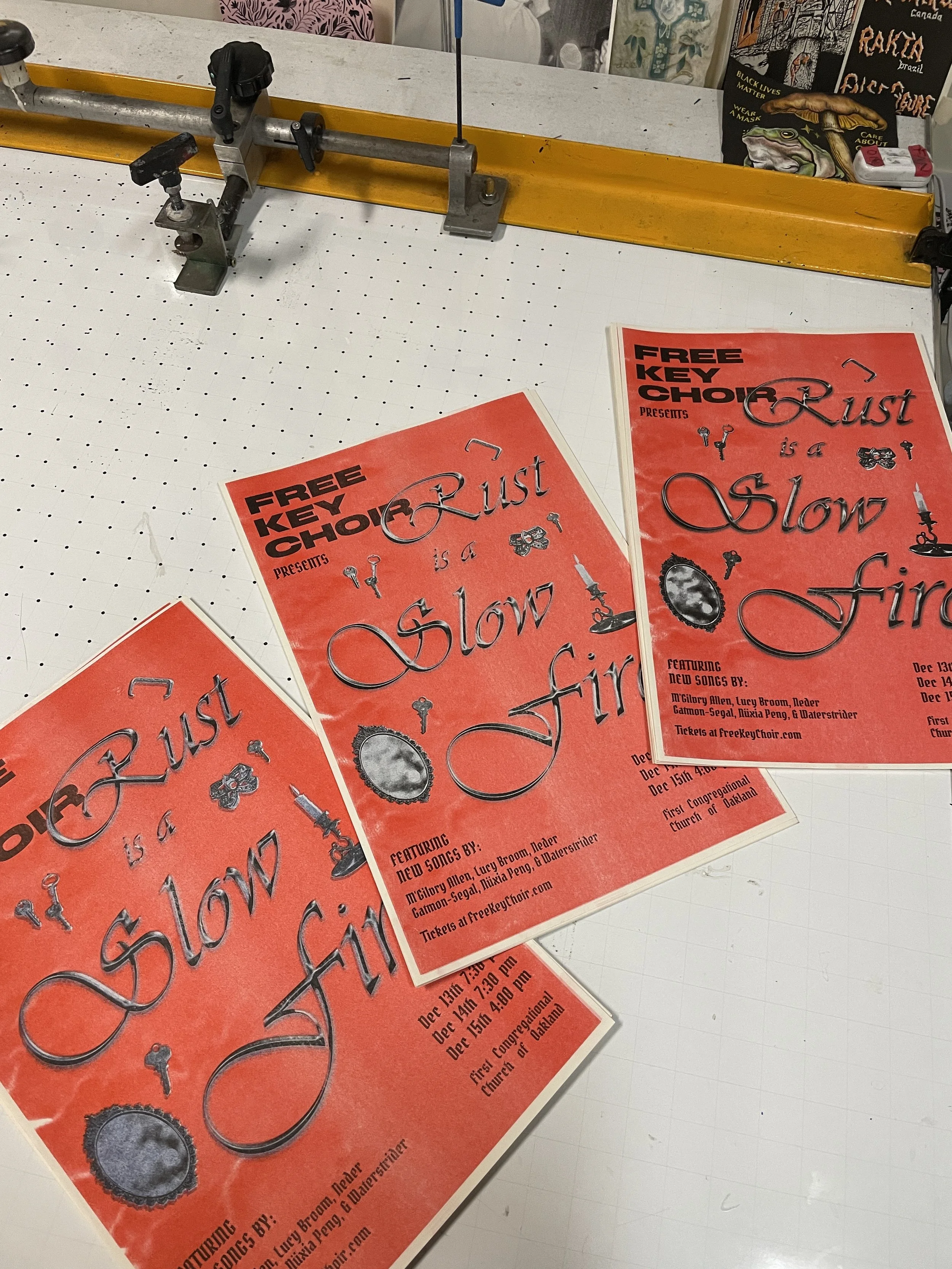 Three red, risograph printed posters advertising a choir concert titled 'Rust is a Slow Fire'. The posters list dates, times, and featured singers. The posters are placed on a cutting mat in a creative workspace.
