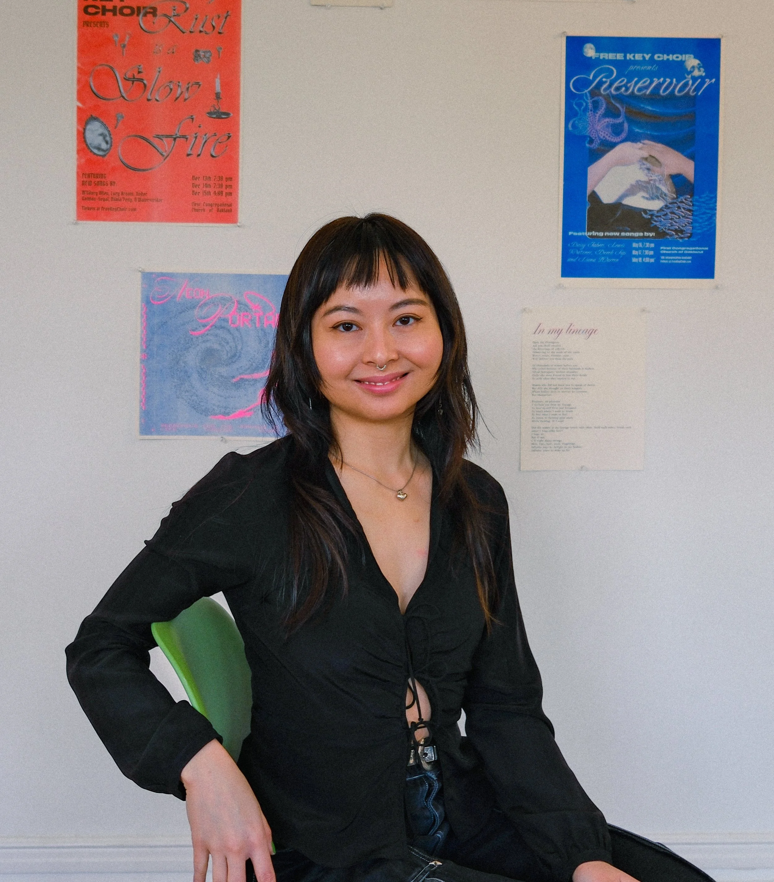 A woman with shoulder-length dark hair and a septum piercing smiling at the camera, seated on a green chair inside a room with posters on the wall behind her.