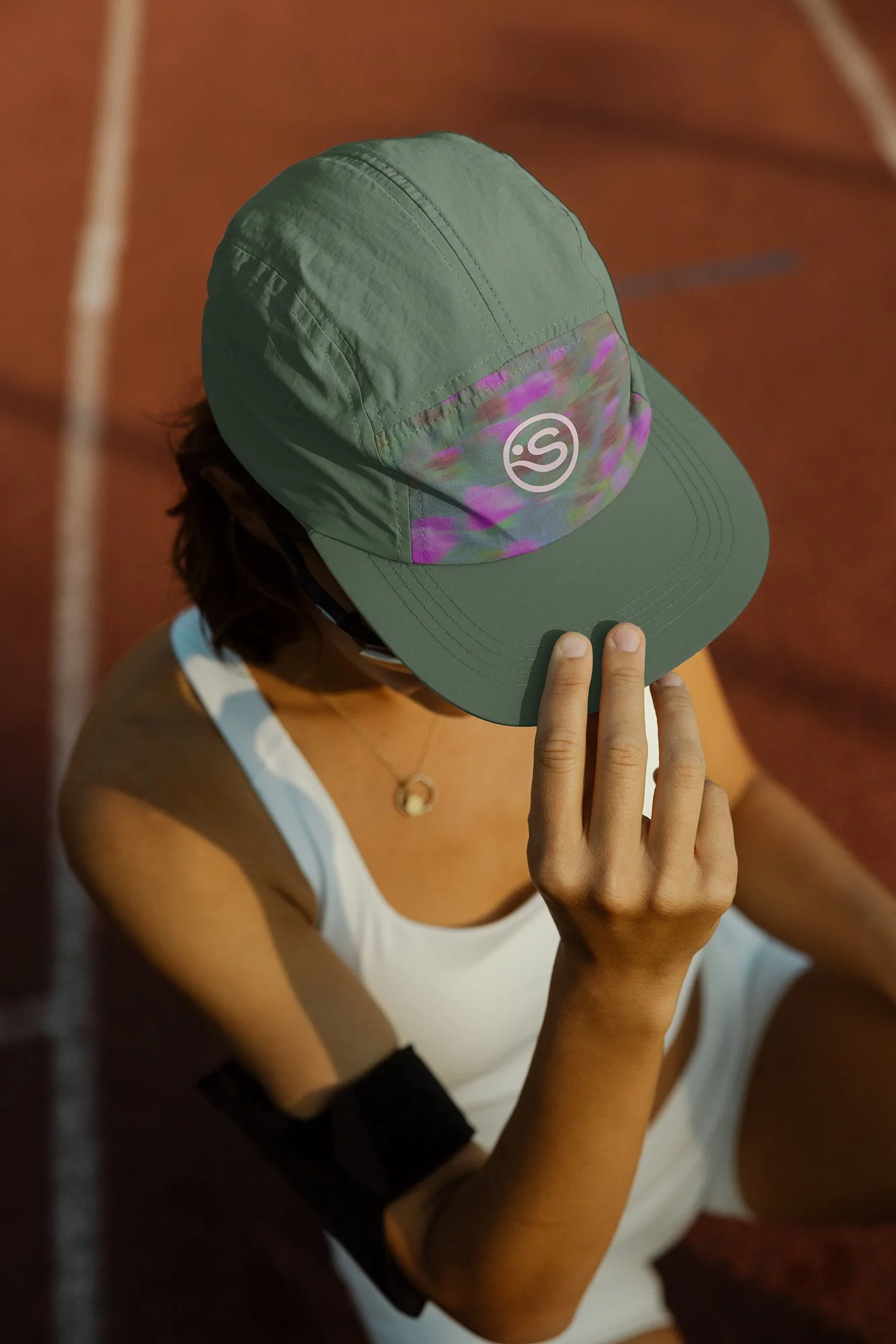A woman sitting on a red outdoor track, holding the brim of a green cap with a logo that looks like the letter "s" on top of a multicolored background of fuchsia, dark red, and subtle blues.