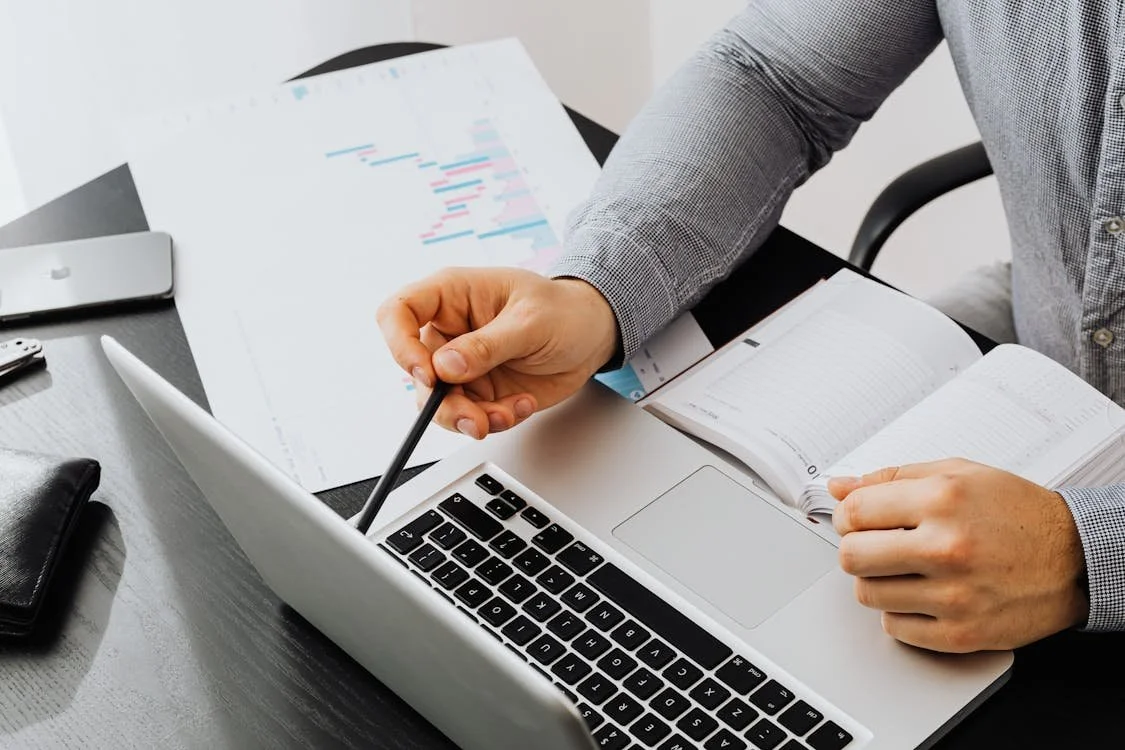 Person pointing at a laptop screen with a pen, surrounded by open notebooks, documents, and a wallet on a desk.