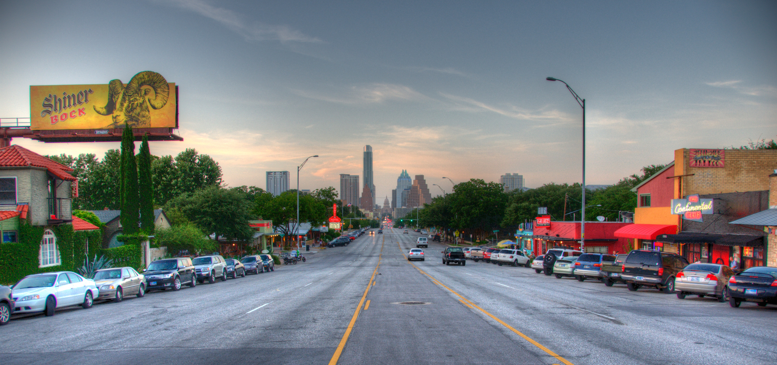 A city street scene at dusk with parked cars along both sides, signs for bars and shops, and skyscrapers in the background under a partly cloudy sky.