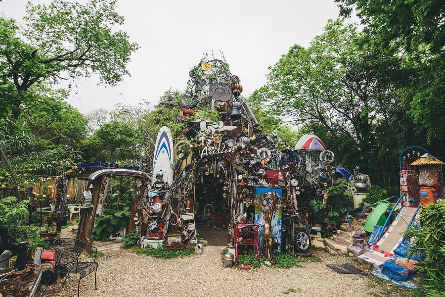 A whimsical playground with an art installation made of recycled materials resembling a mountain or fairy house, surrounded by trees and playful structures.