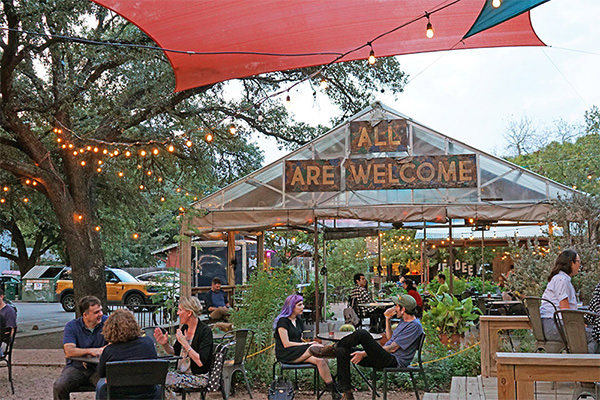 Outdoor gathering space with string lights and a glasshouse with a welcoming sign, surrounded by people sitting and socializing.