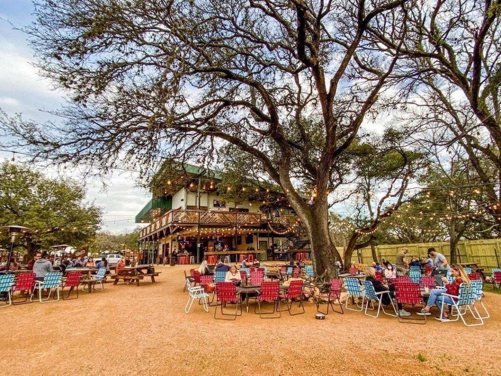 Outdoor dining area with colorful table and chair arrangements, a large tree at the center, string lights, and a two-story building in the background, during daytime.