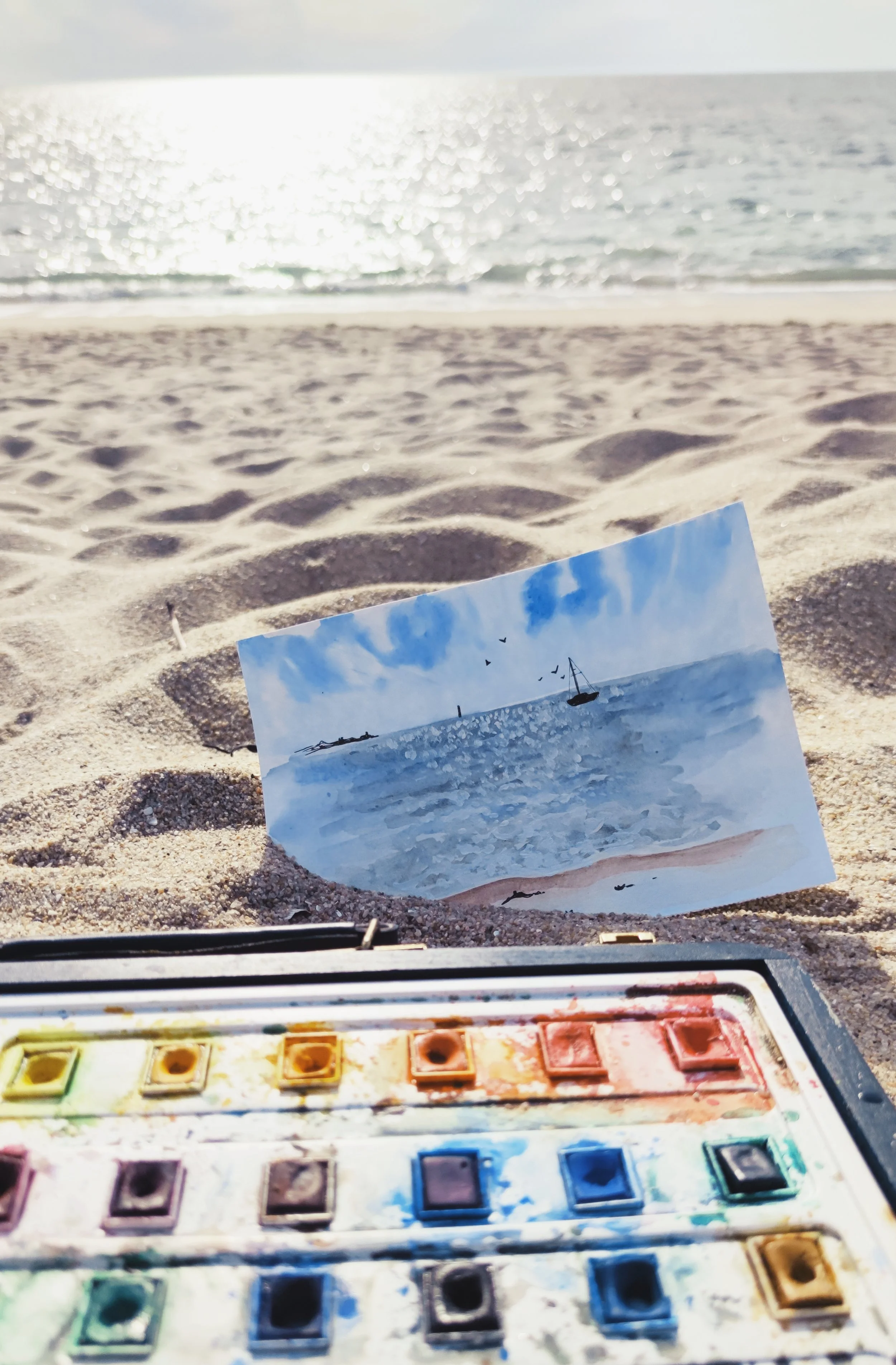 A photo of a sandy beach with the sea in the background, a watercolor paint set in the foreground, and a photograph of a seascape propped up in the sand.