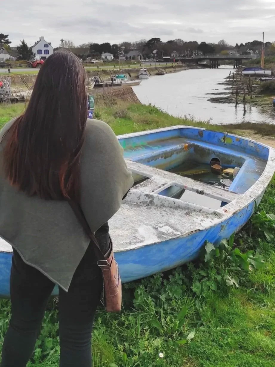 Une femme regarde un bateau abandonné à terre près d'une rivière, dans une zone verdoyante et résidentielle.